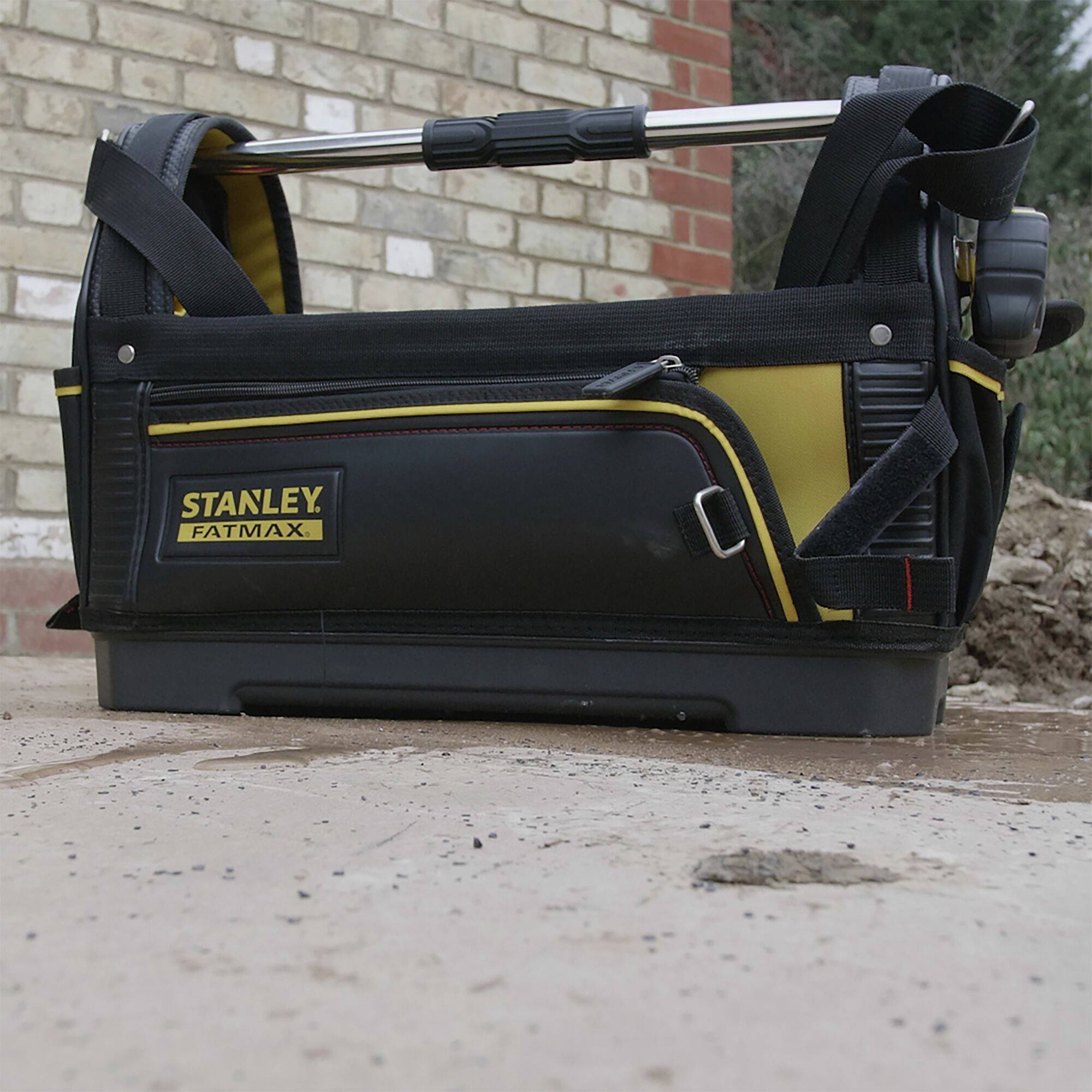 A black and yellow Stanley tool bag with carrying straps and tools, placed on a paved surface in front of a brick wall.