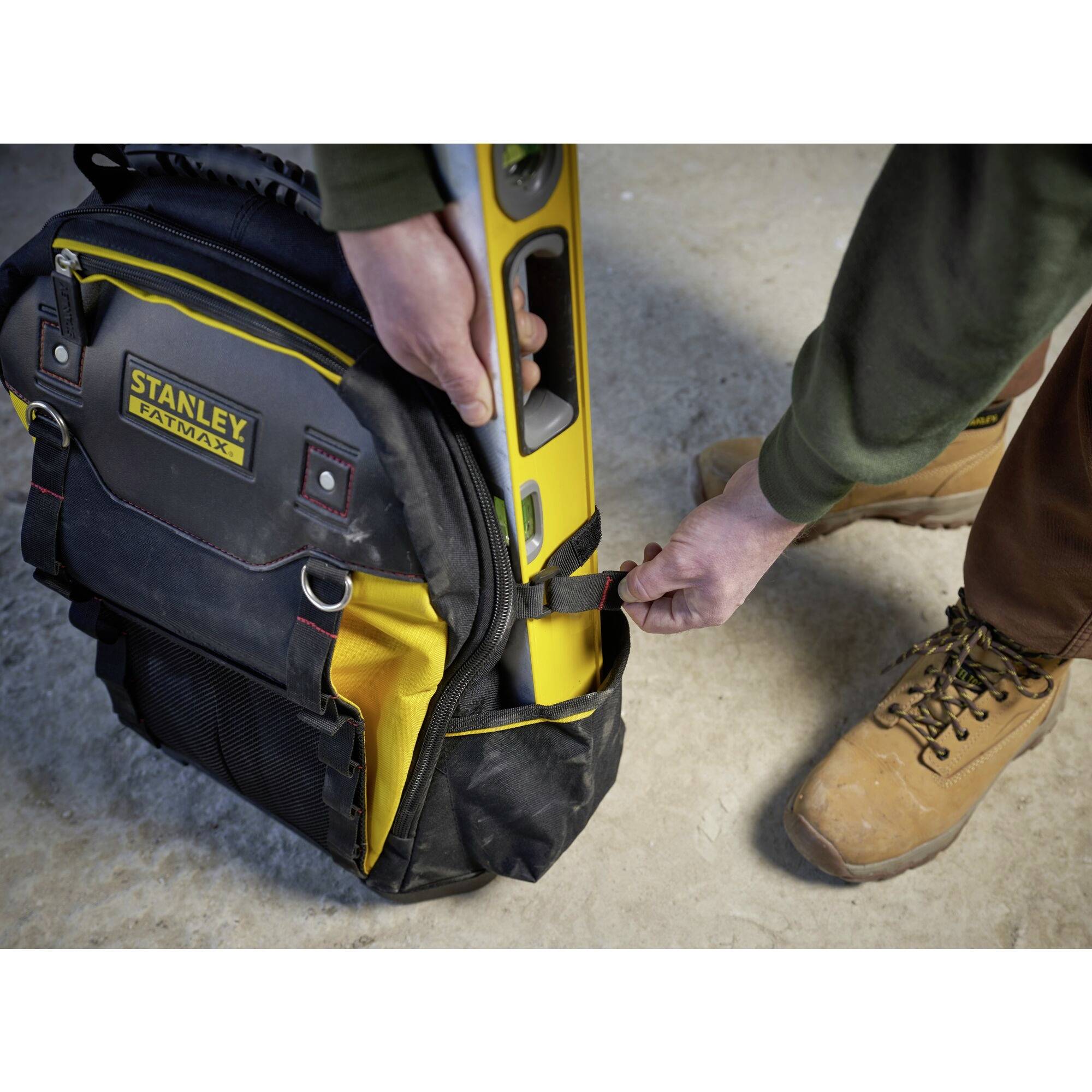 A person is holding a Stanley bag with a spirit level set. The bag is resting on a roughly textured surface, and the person is wearing sturdy boots.