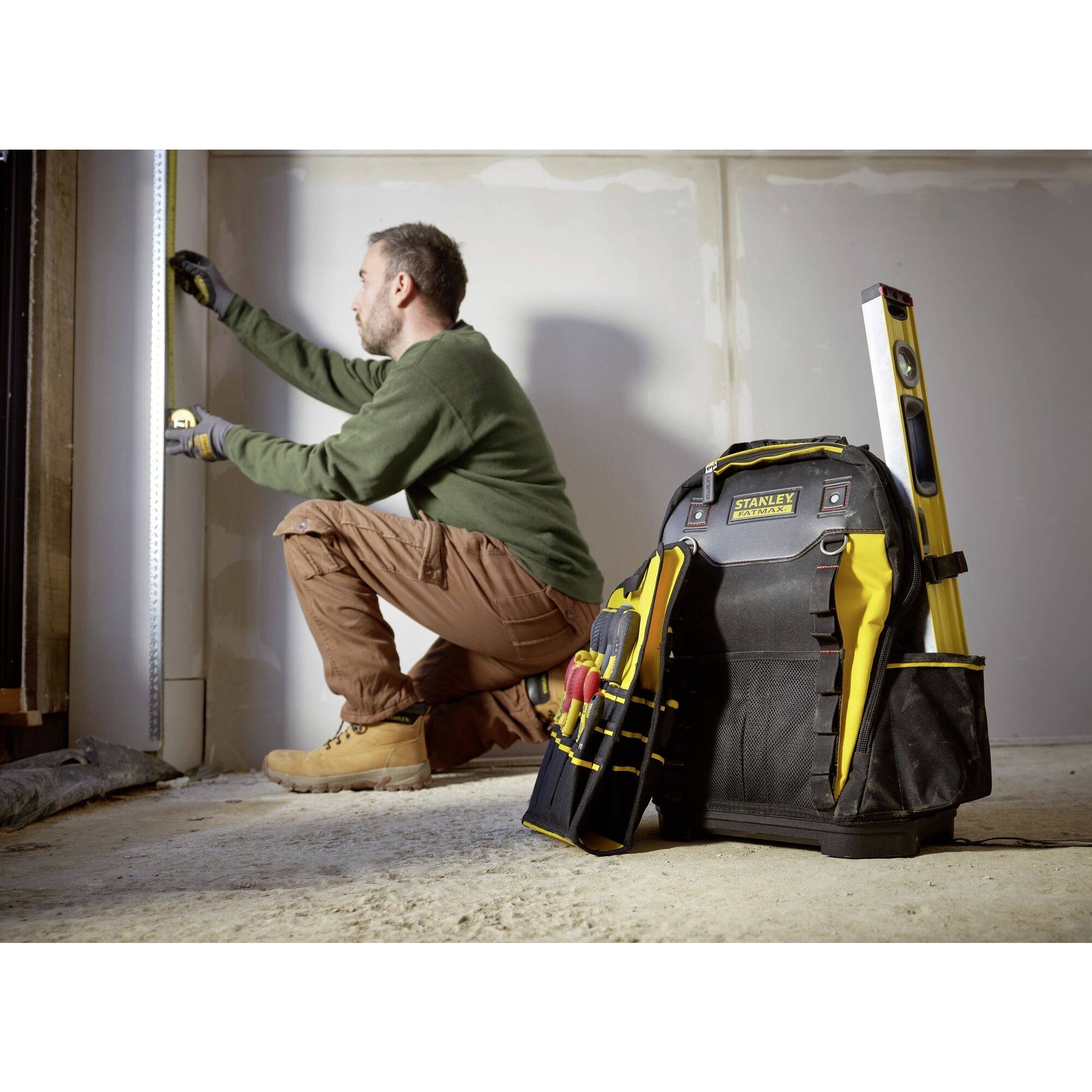 A man is measuring a wall with a tape measure, kneeling on the floor. Next to him is a tool backpack with various tools.