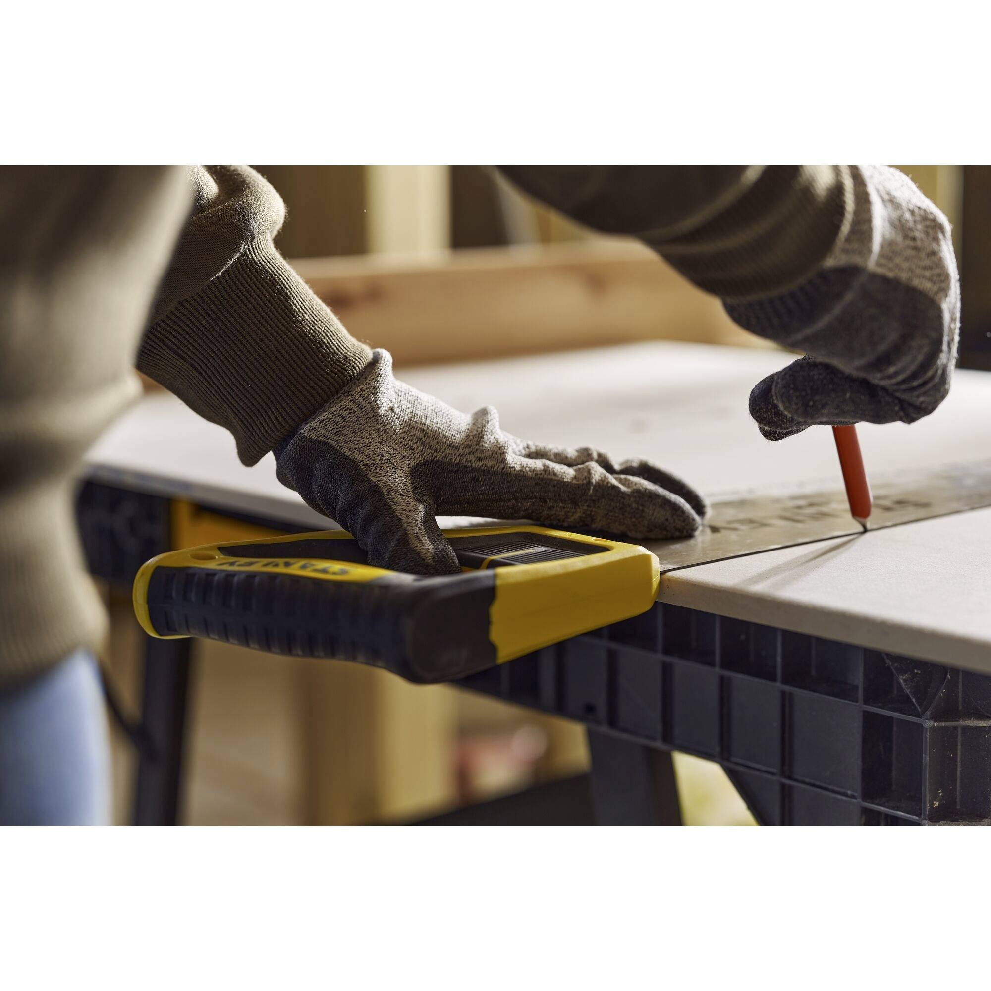 A person is wearing work gloves, using a handsaw and marking a wooden board with a red pencil on a workbench.