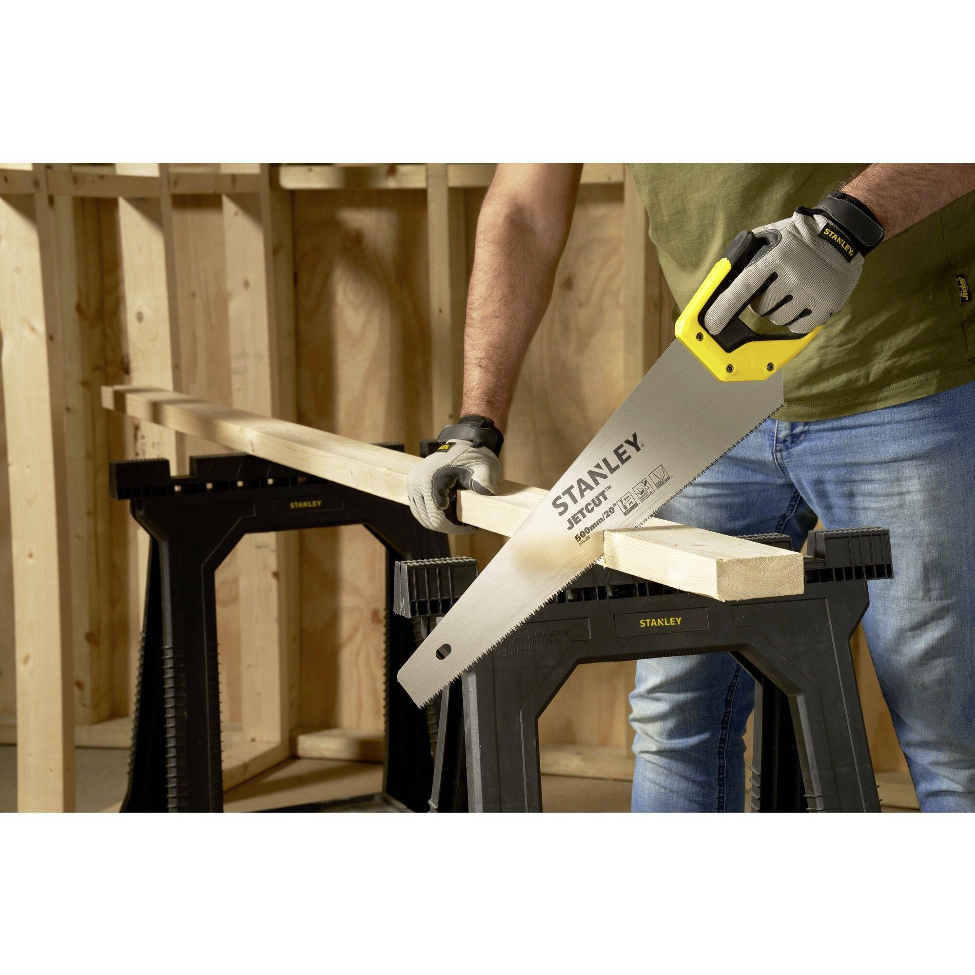 A person is sawing a wooden board on two sawhorses using a Stanley handsaw. The person is wearing gloves and jeans.