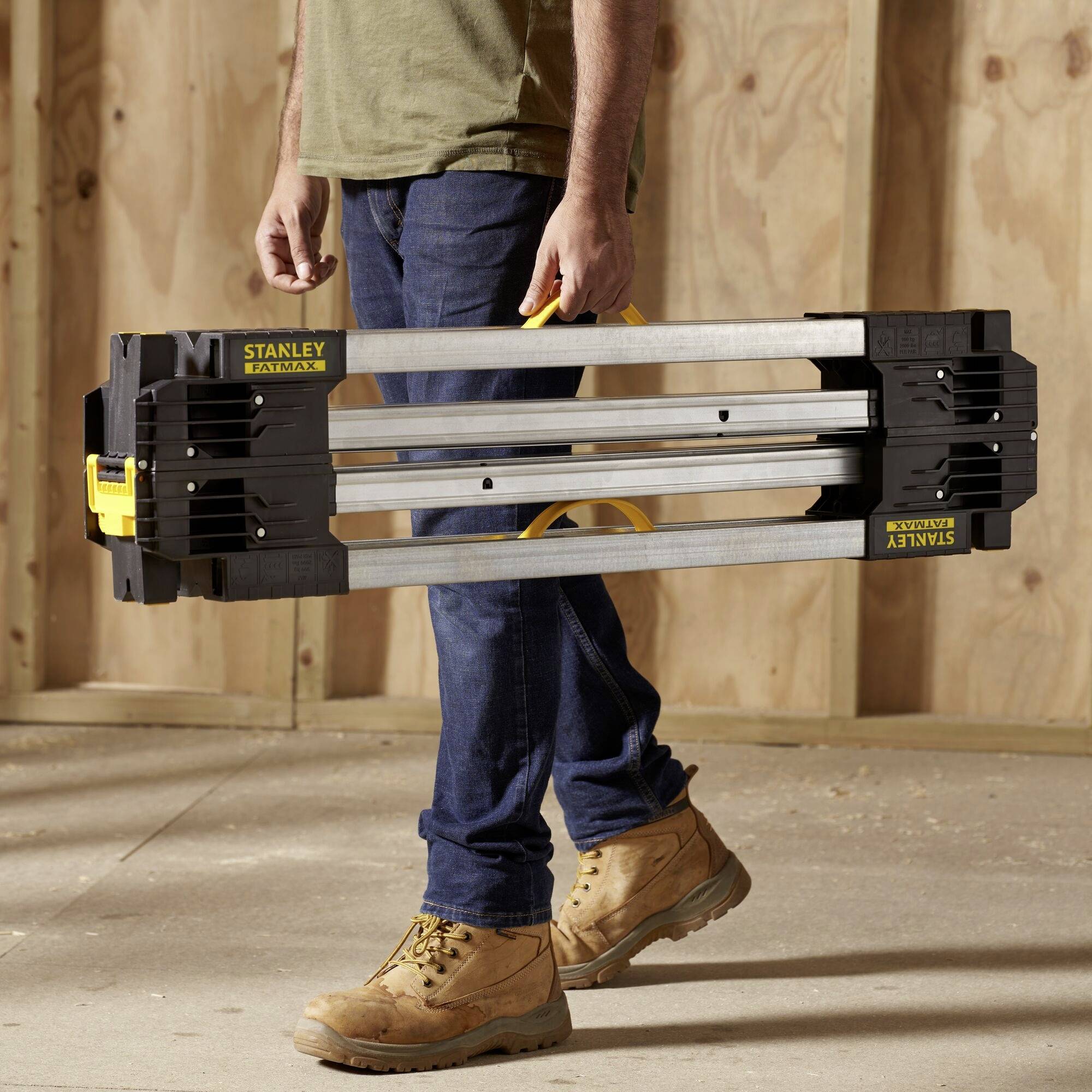 A person in workwear is carrying a folded building material holder in an unfinished room with wooden walls.