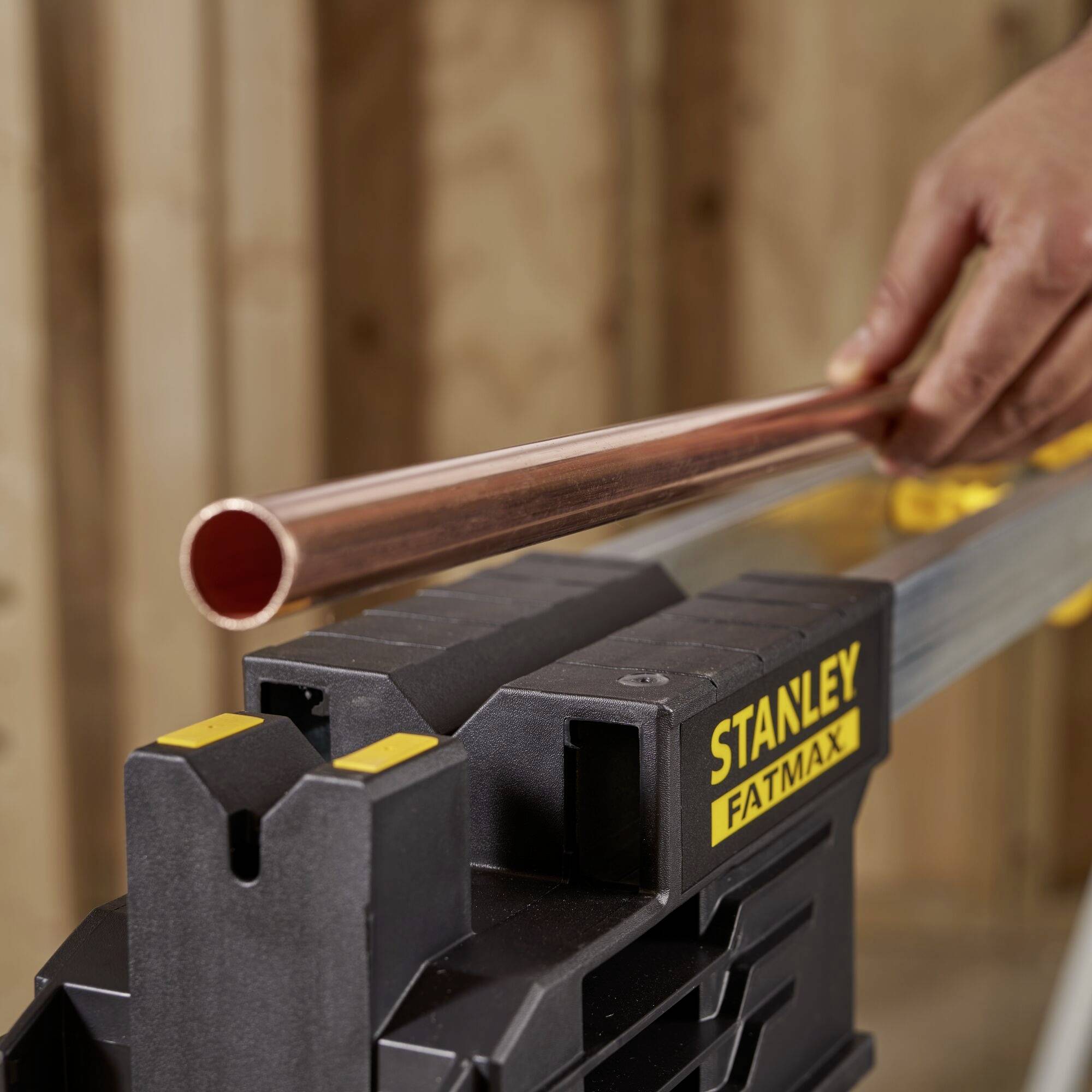 A hand is guiding a copper pipe over a yellow pipe cutting machine branded 'Stanley Fatmax' in a workshop environment.