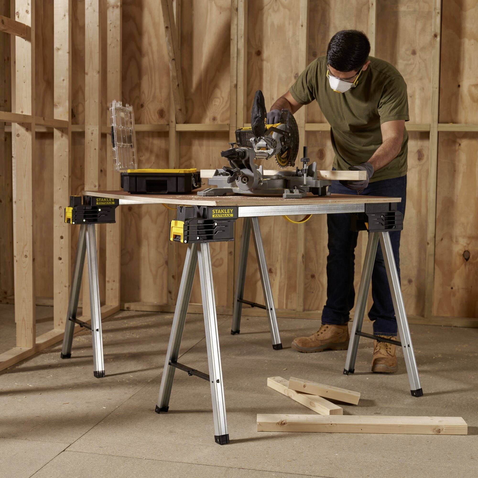 A man is cutting wood with a circular saw in an unfinished room. He is wearing a mask and standing at a workbench.