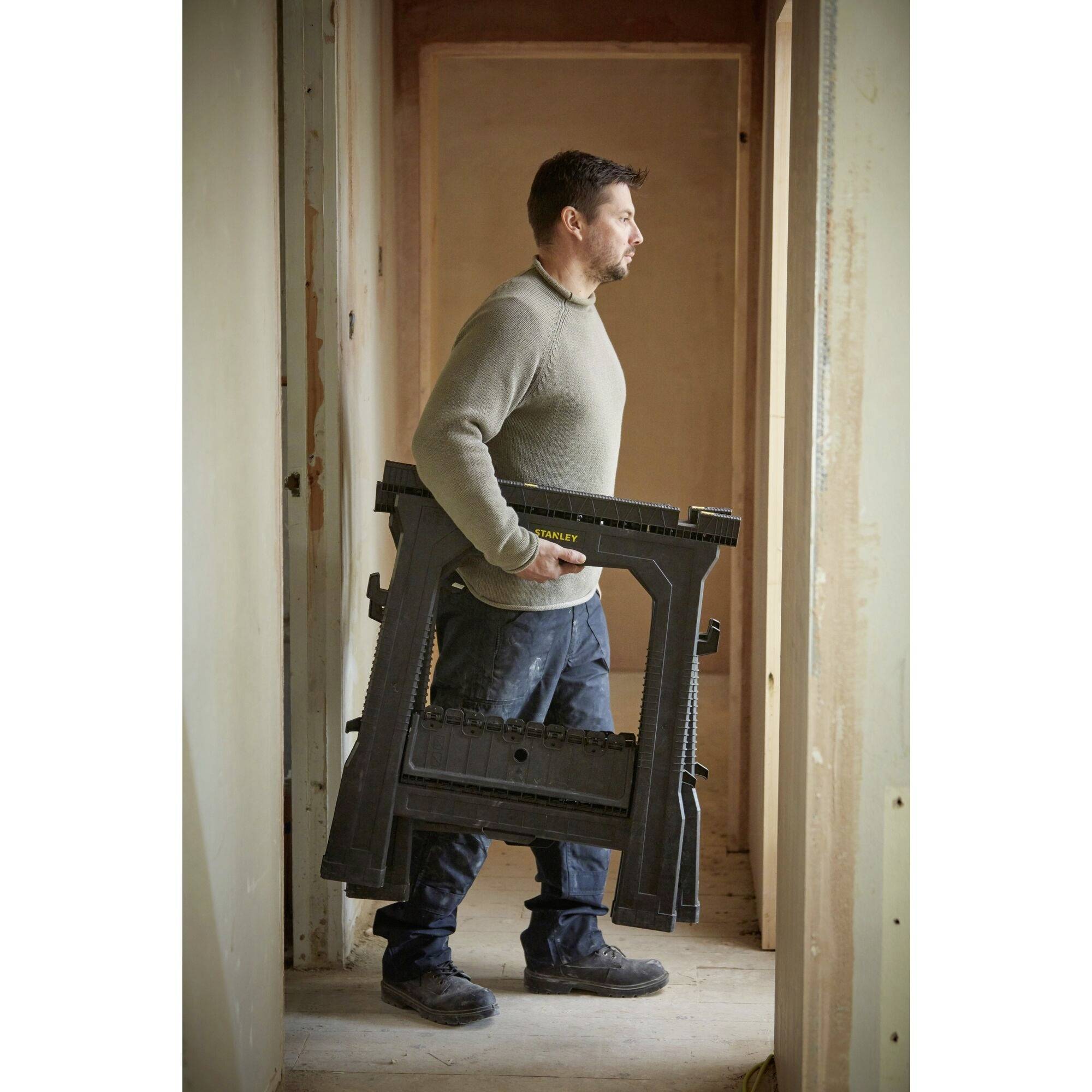 A man in workwear is carrying a foldable black workbench in an unfinished room. He is looking outside.