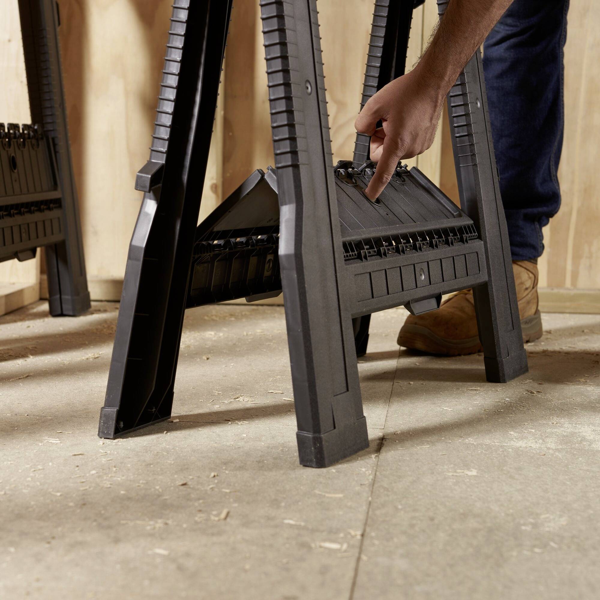 A person is positioning a black, folding sawbuck frame on a wooden floor, preparing it for use.