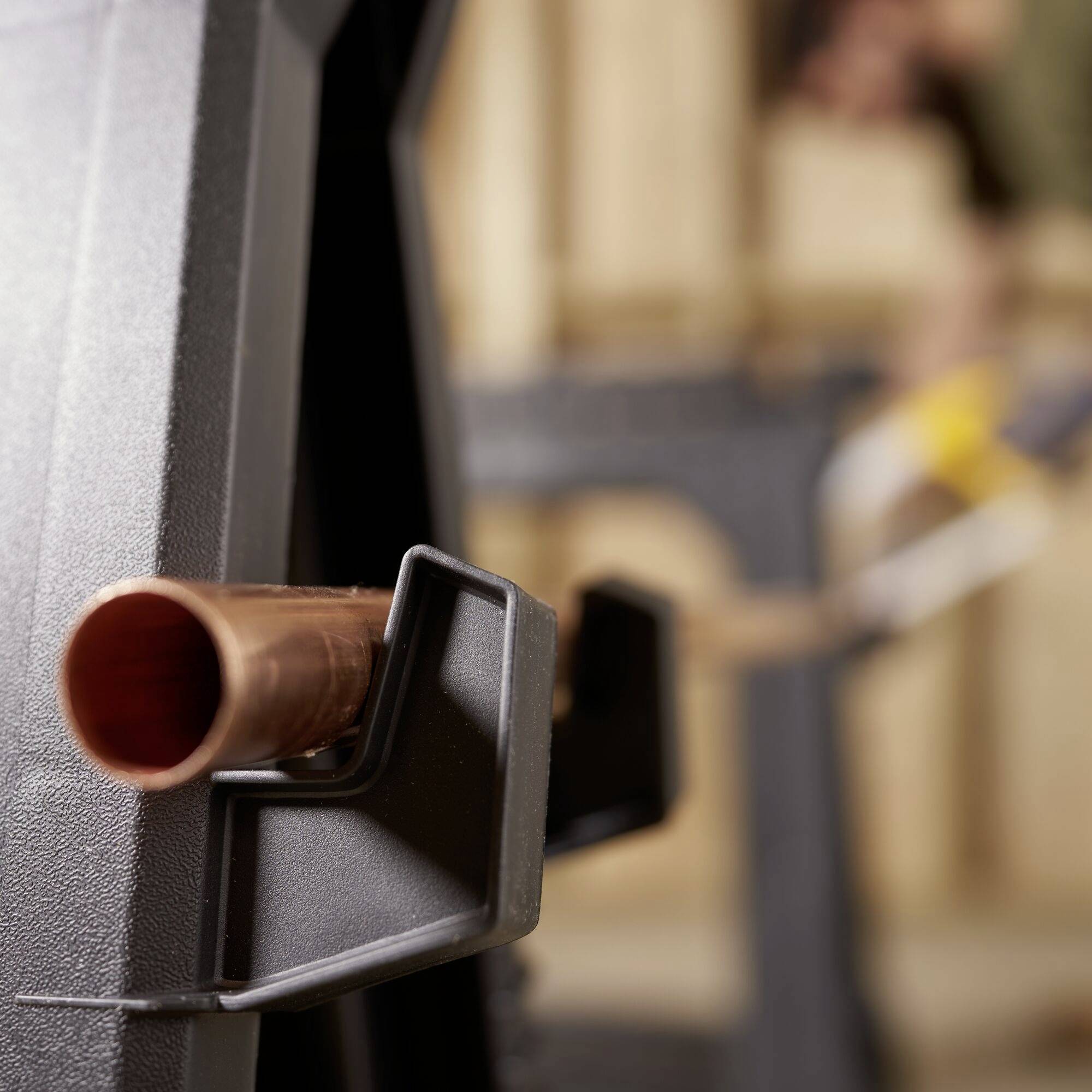 Close-up of a copper pipe opening secured in a clamp, with a blurred woodworking scene in the background.