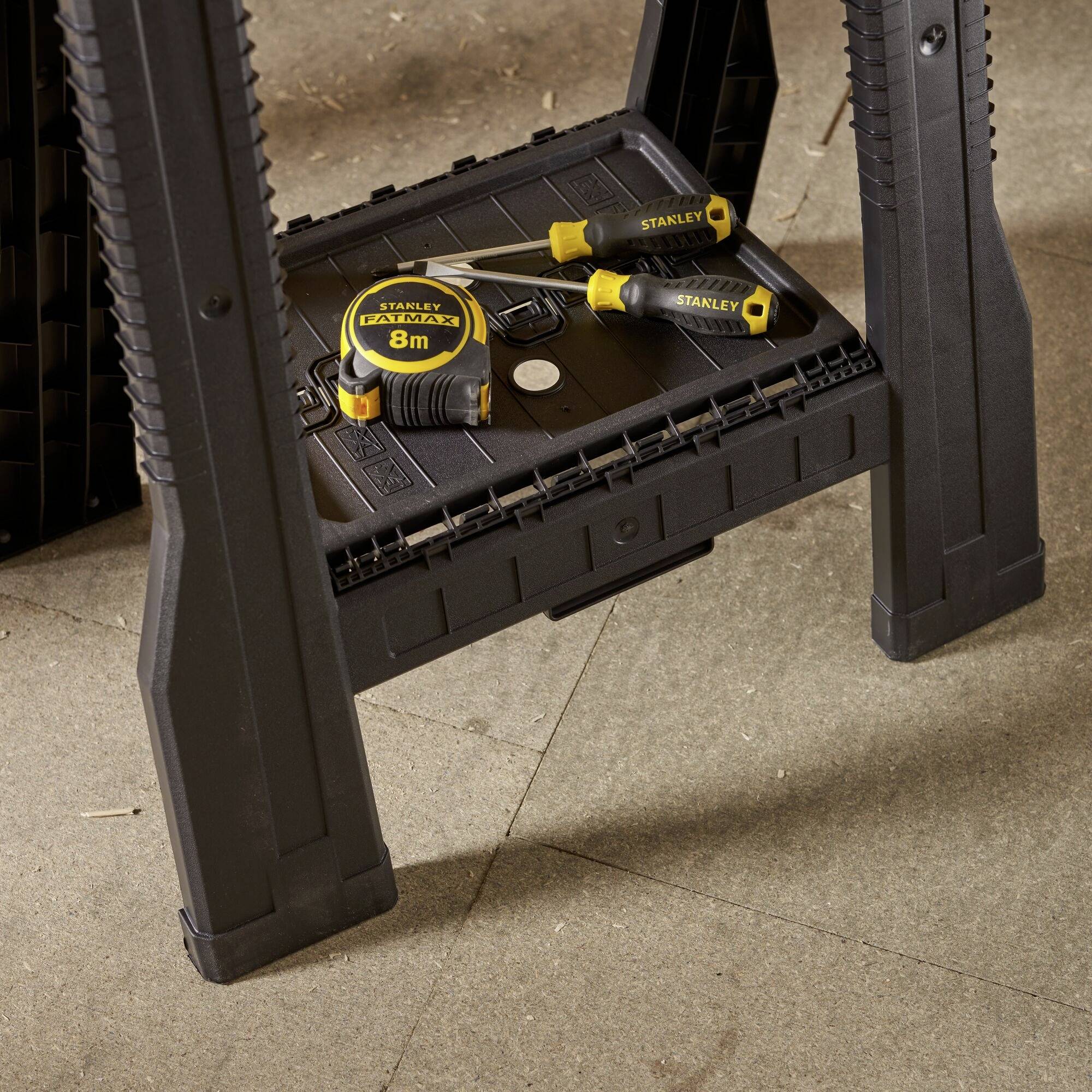 Black folding step stool with yellow tape measure and screwdrivers placed on it; 'STANLEY' tool branding visible on all tools.
