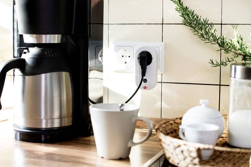 Coffee machine on worktop, plugged into wall socket, with a cup and teapot beside it in the bright kitchen area.