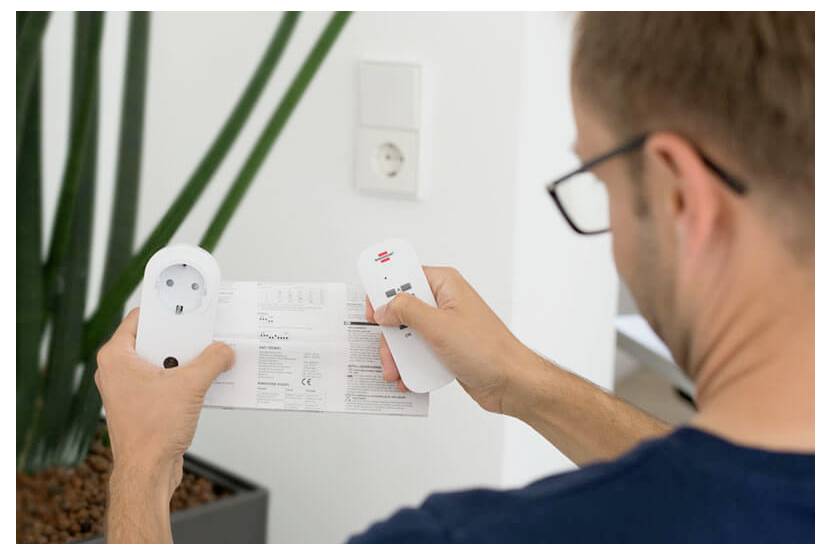 Man holding a remote control and instruction sheet while examining a wall outlet or smart plug during setup.