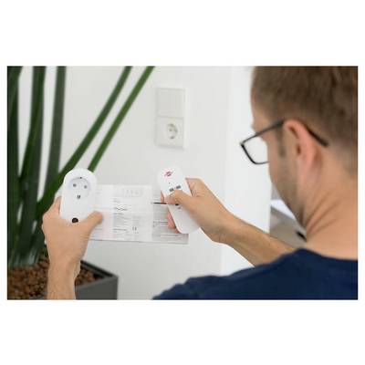 Man holding a remote control and instruction sheet while examining a wall outlet or smart plug during setup.