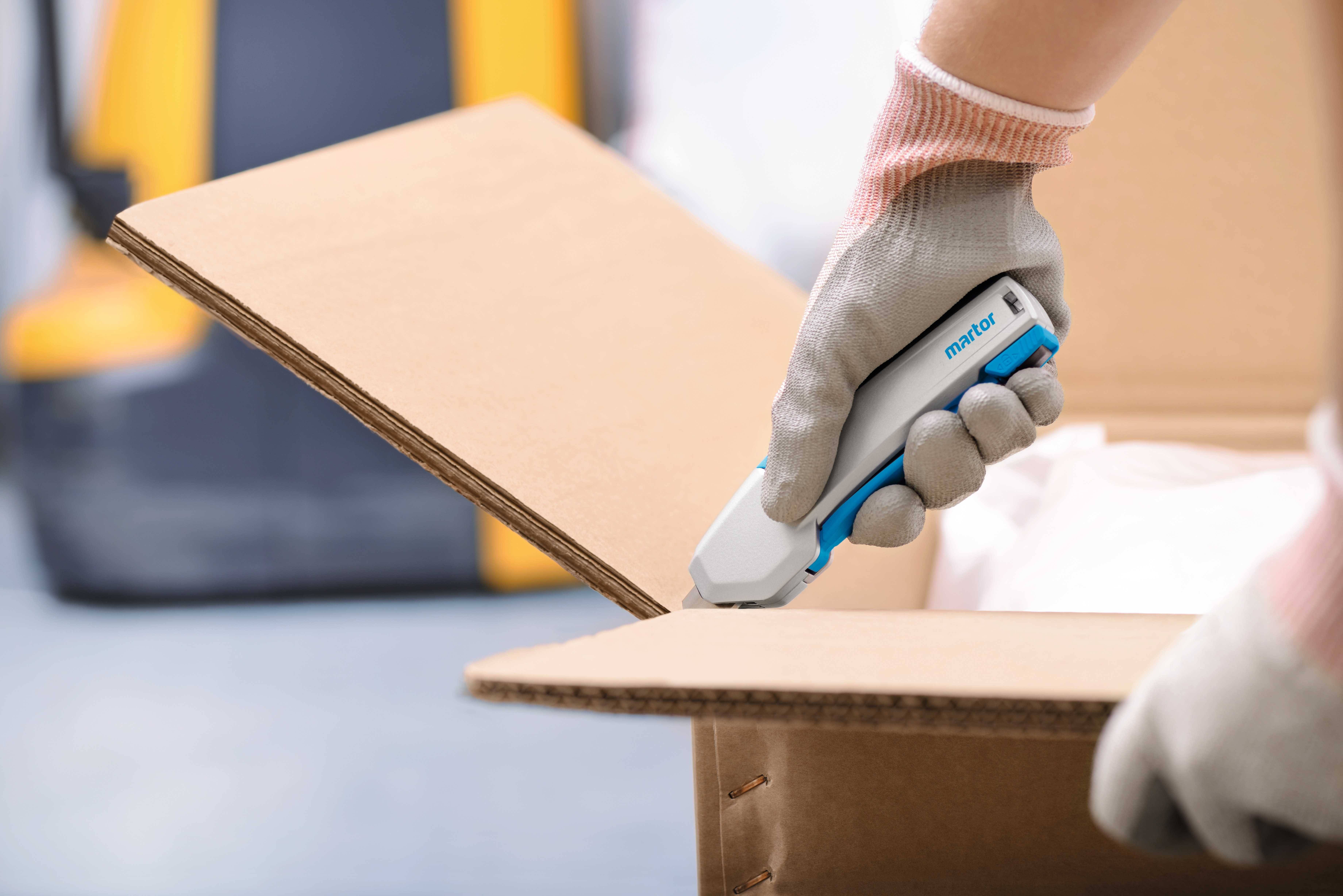 A person opens a cardboard box using a safety box cutter. A forklift is visible in the blurred background.