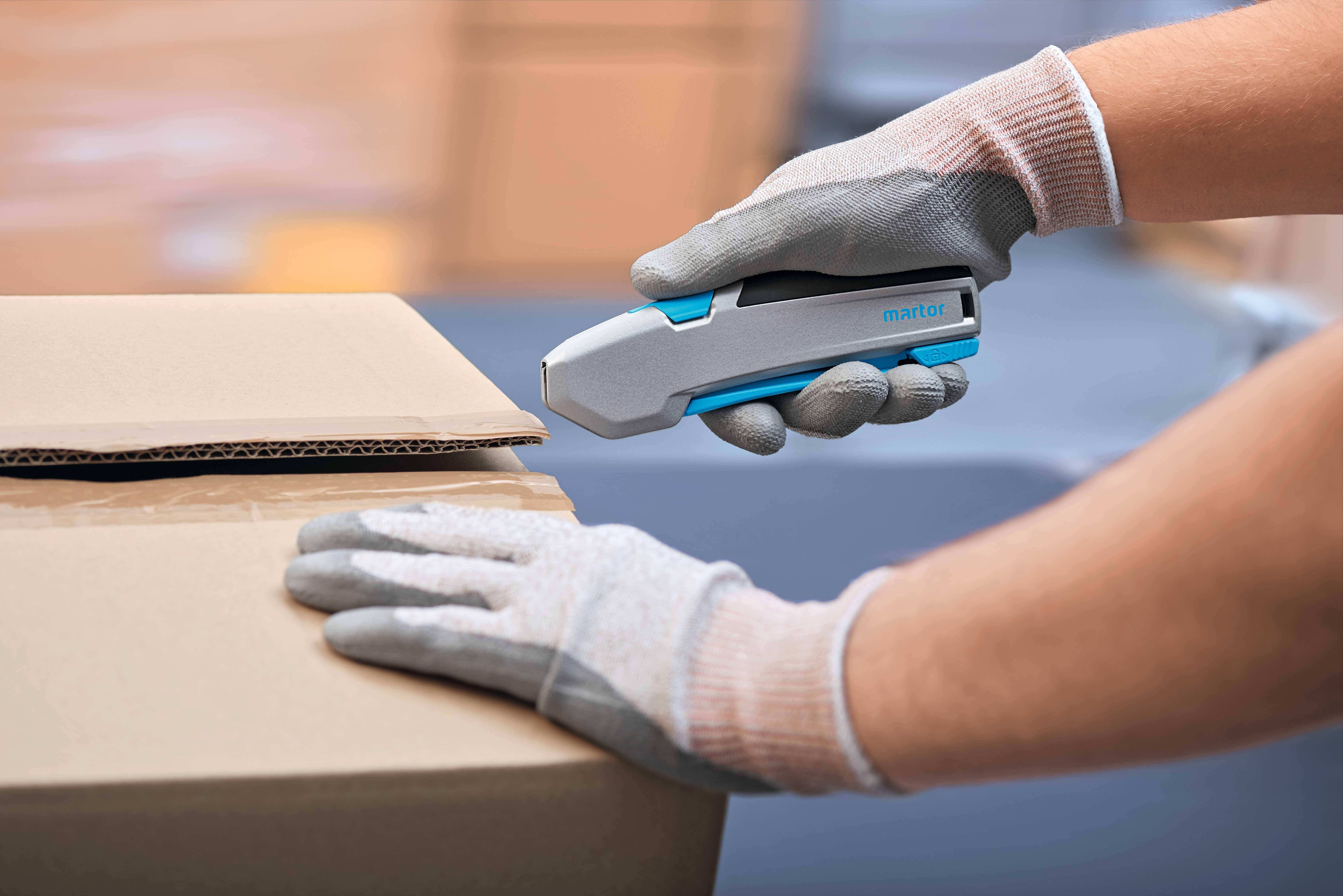 A person wearing gloves carefully opens a cardboard box containing a grey cutting tool. Background out of focus.