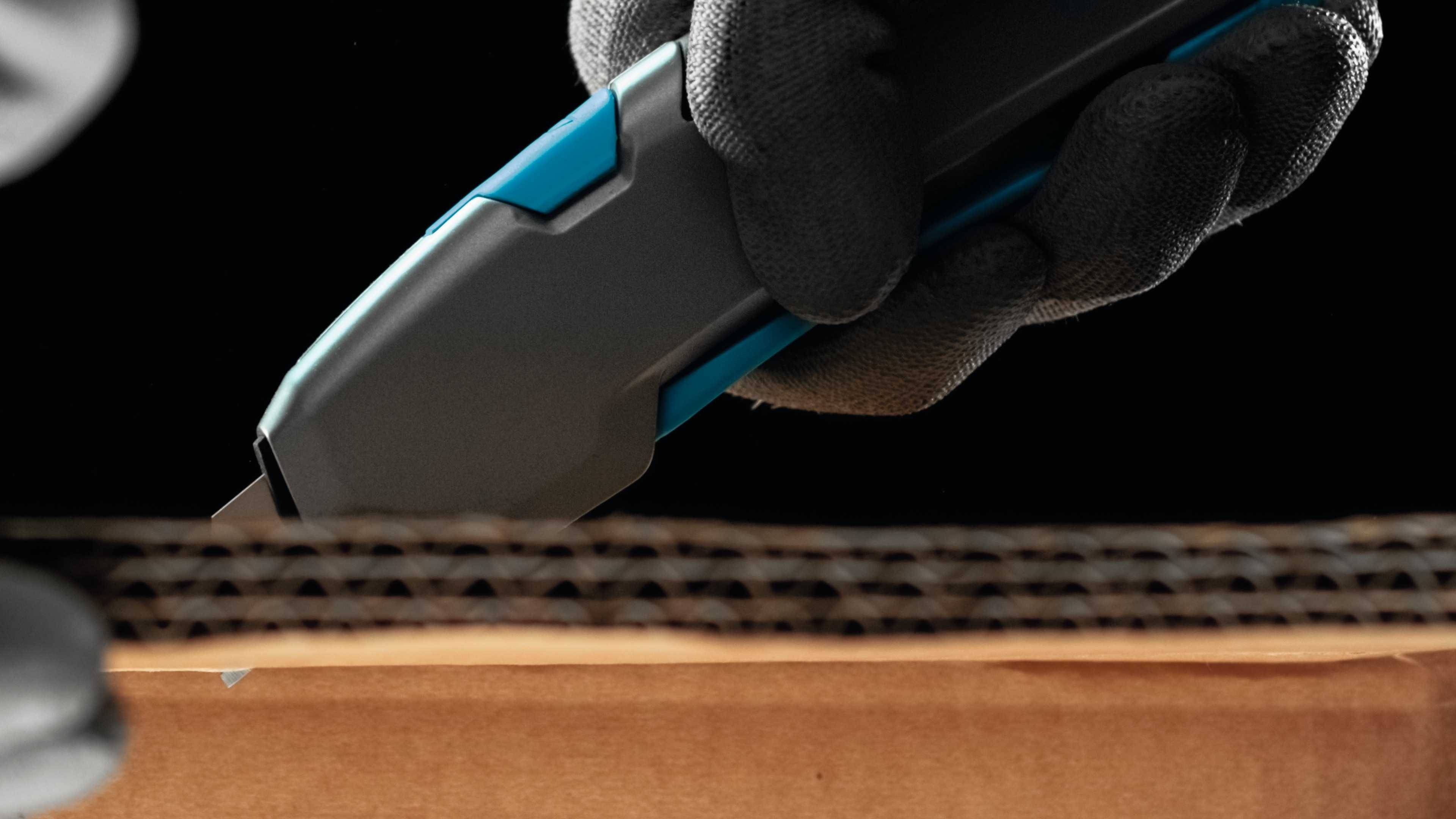 A hand in a glove cuts a cardboard box using a grey and blue craft knife.