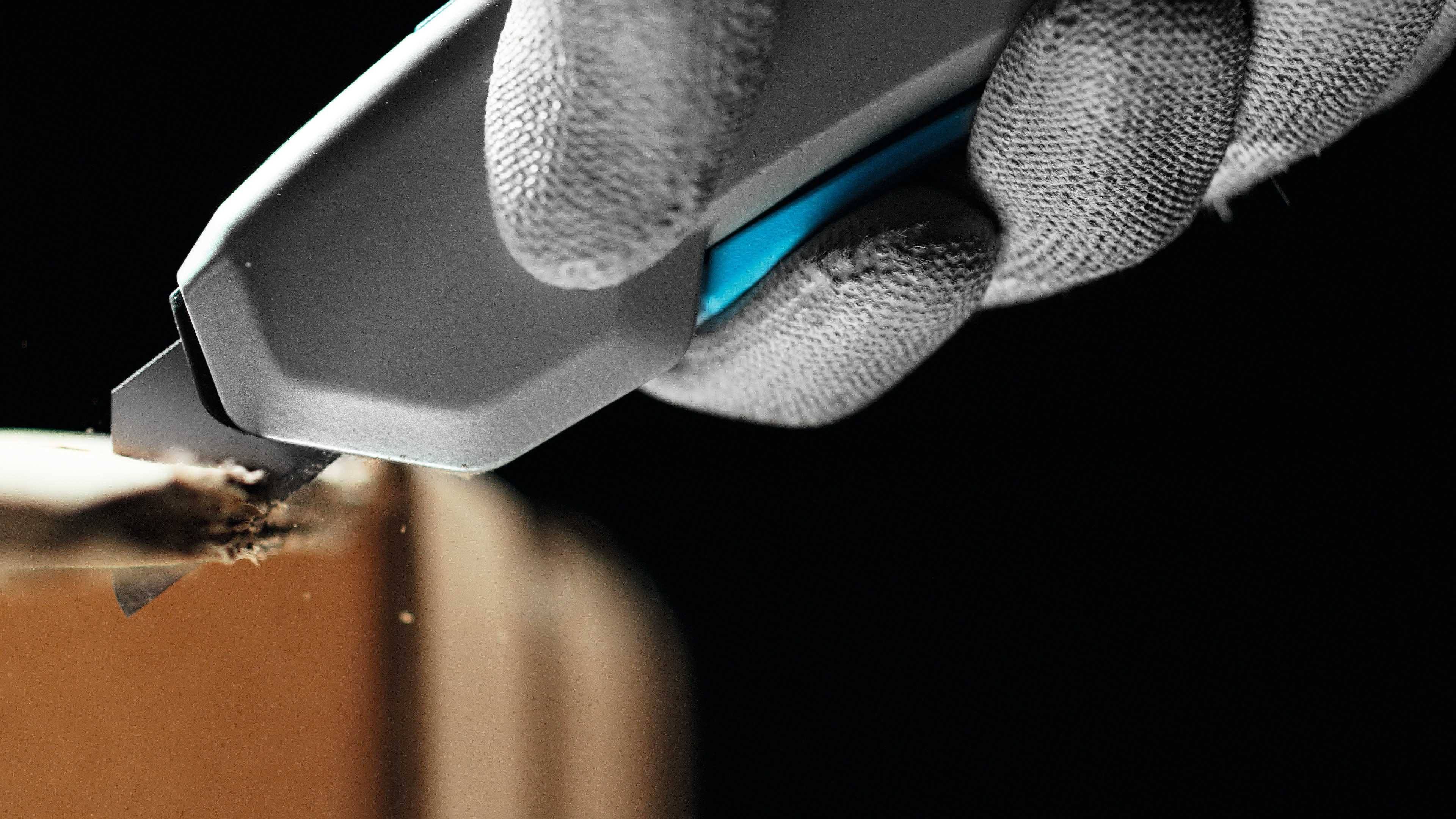 A gloved hand cuts through cardboard with a grey and blue craft knife.