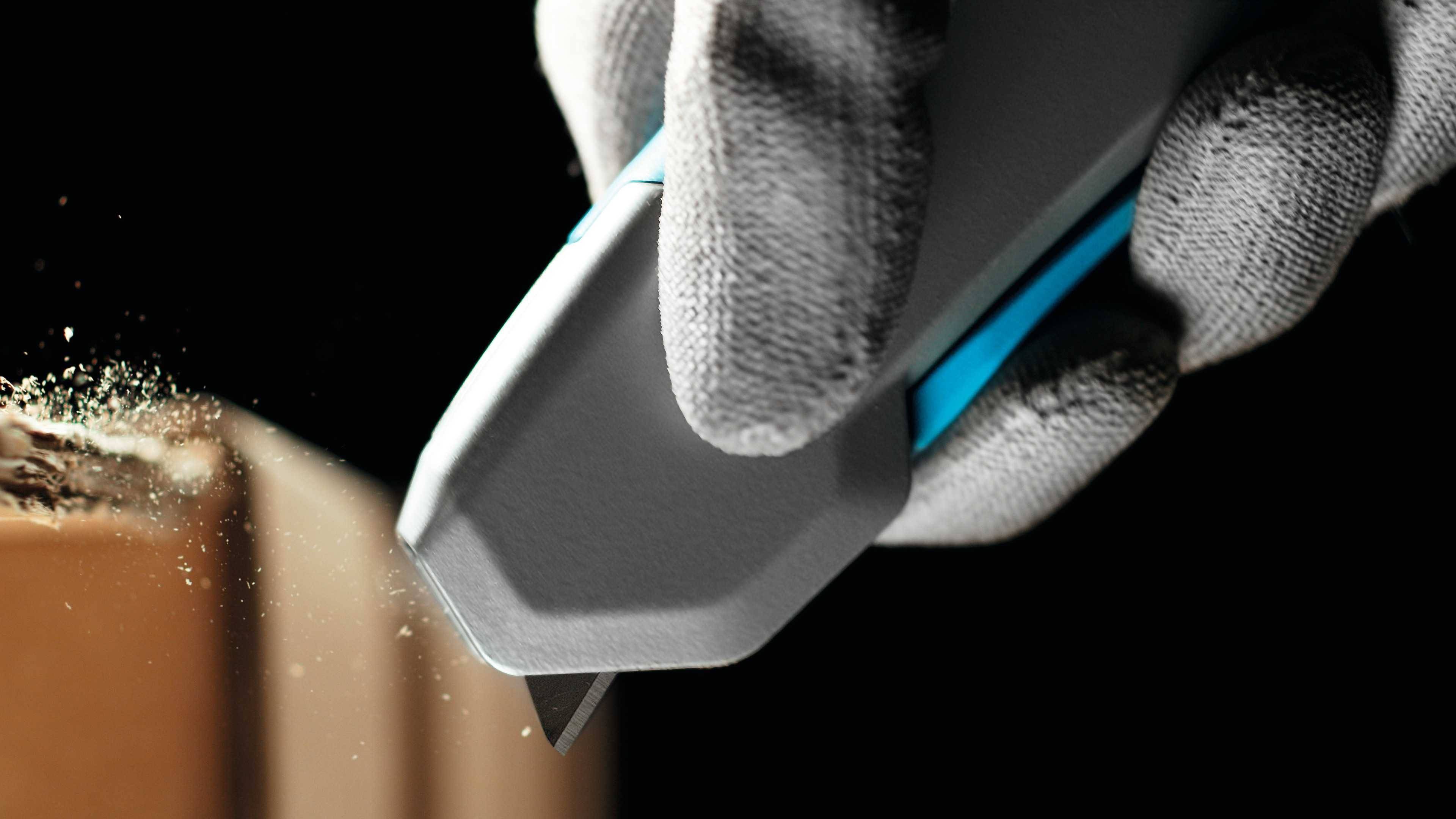 A hand wearing a grey glove holds a craft knife and cuts precisely into a piece of cardboard, with wood shavings visible.