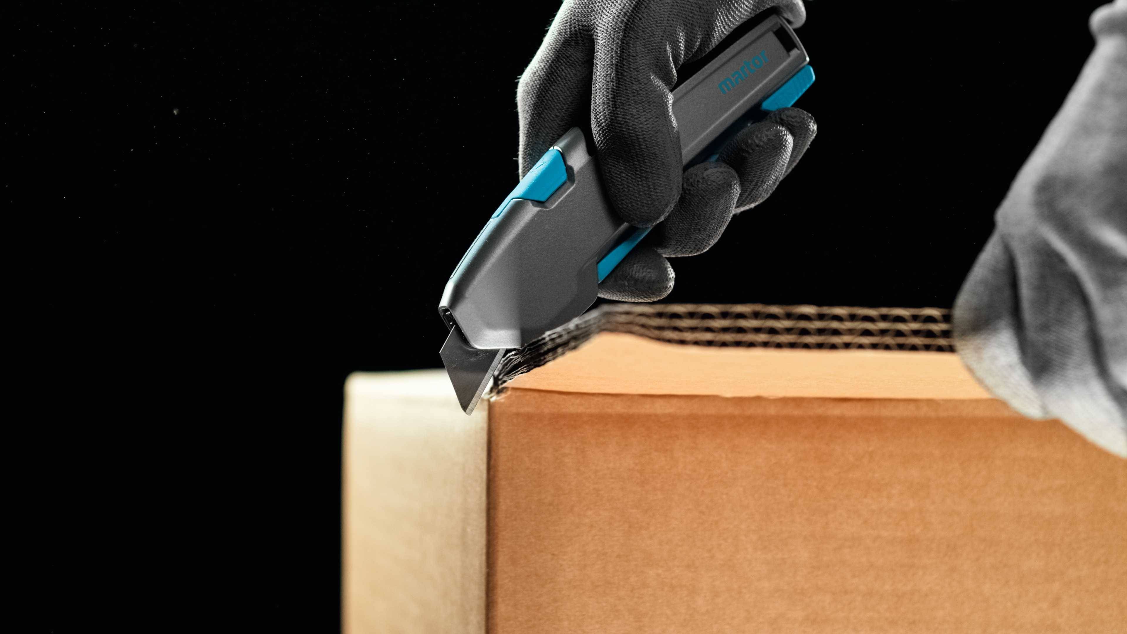 A gloved hand cuts open a cardboard box using a utility knife blade.