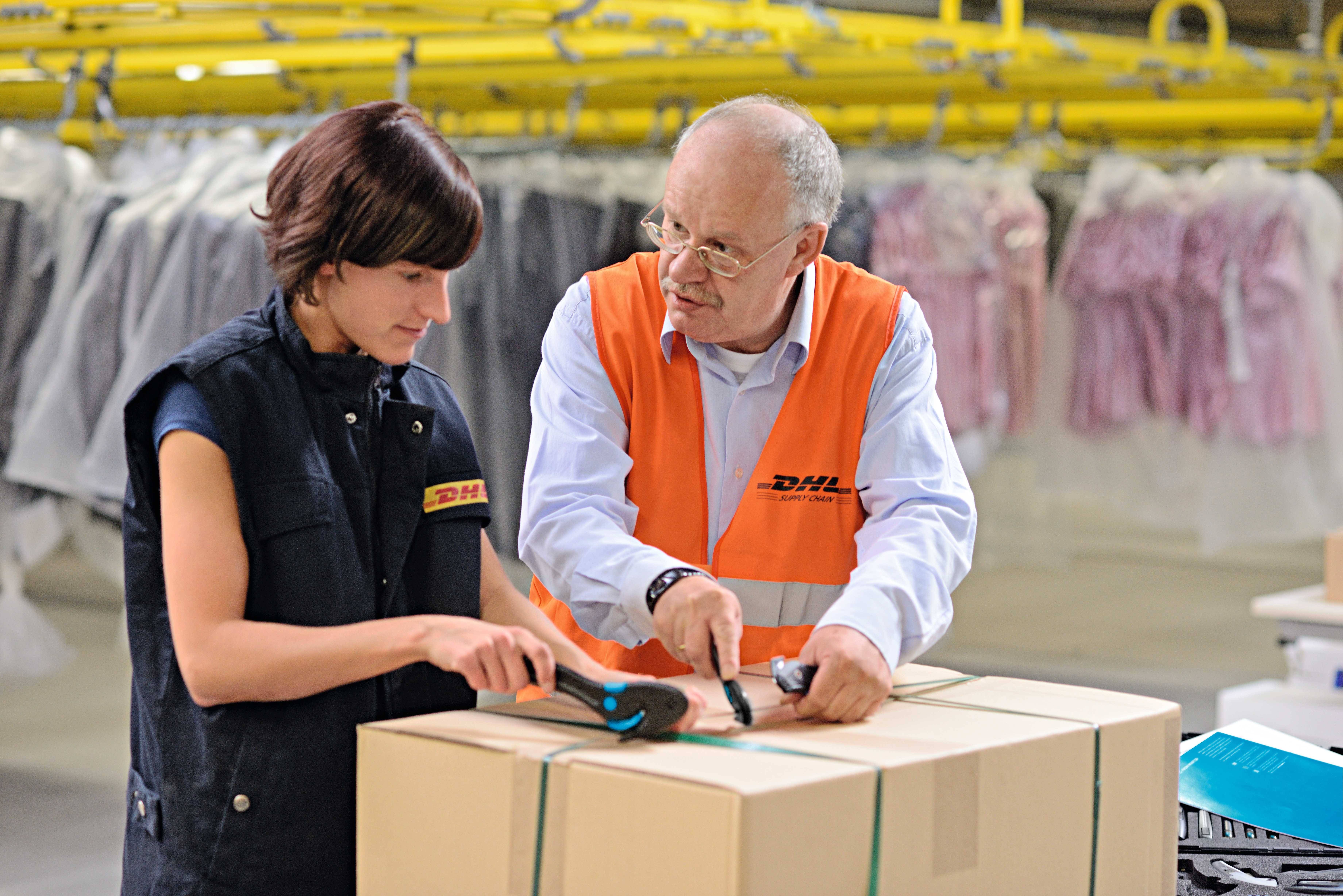 Two people in DHL uniforms are working together on a package in a warehouse. Clothing items can be seen in the background.