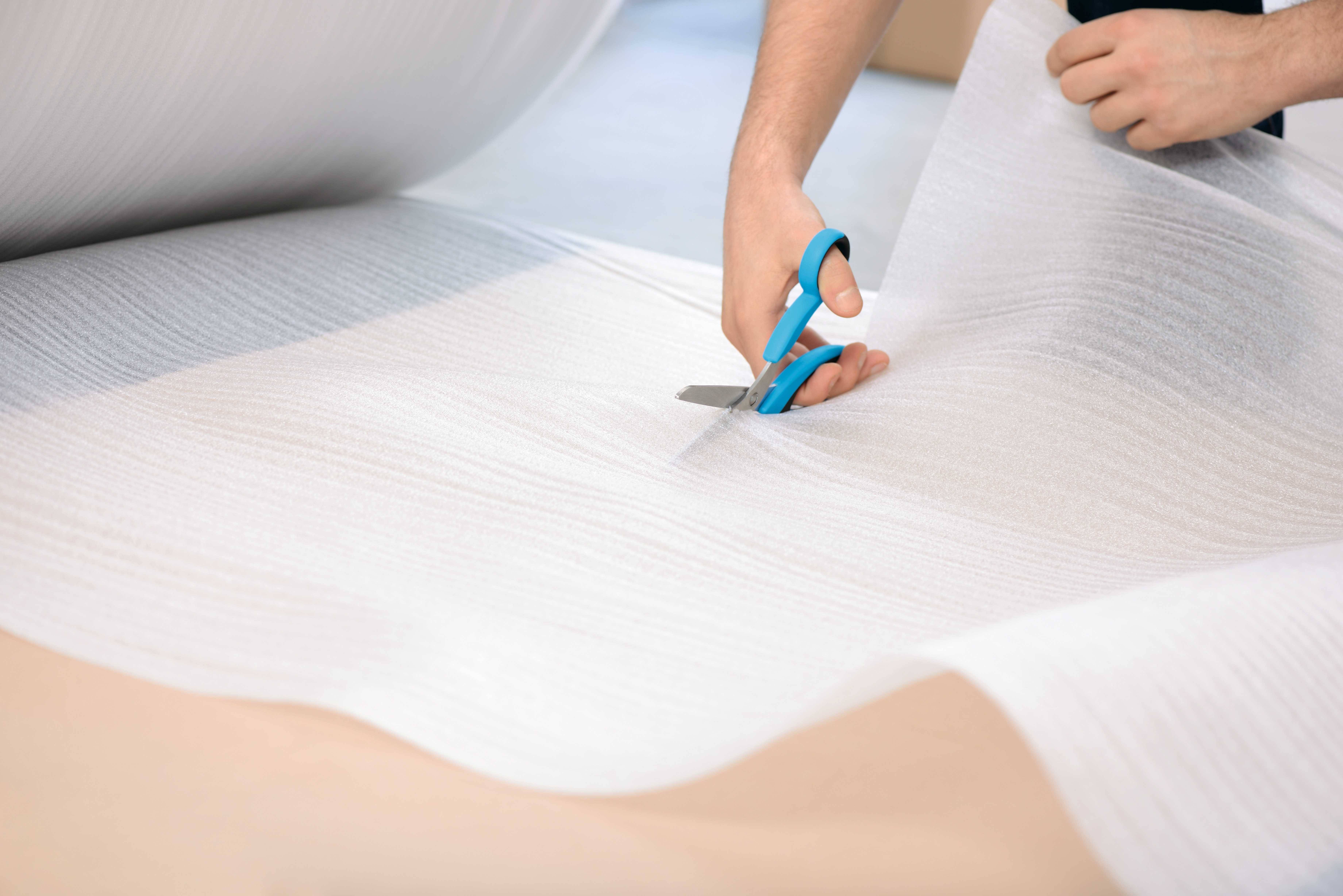 A person is cutting a large sheet of white packaging foam with blue scissors on a table.