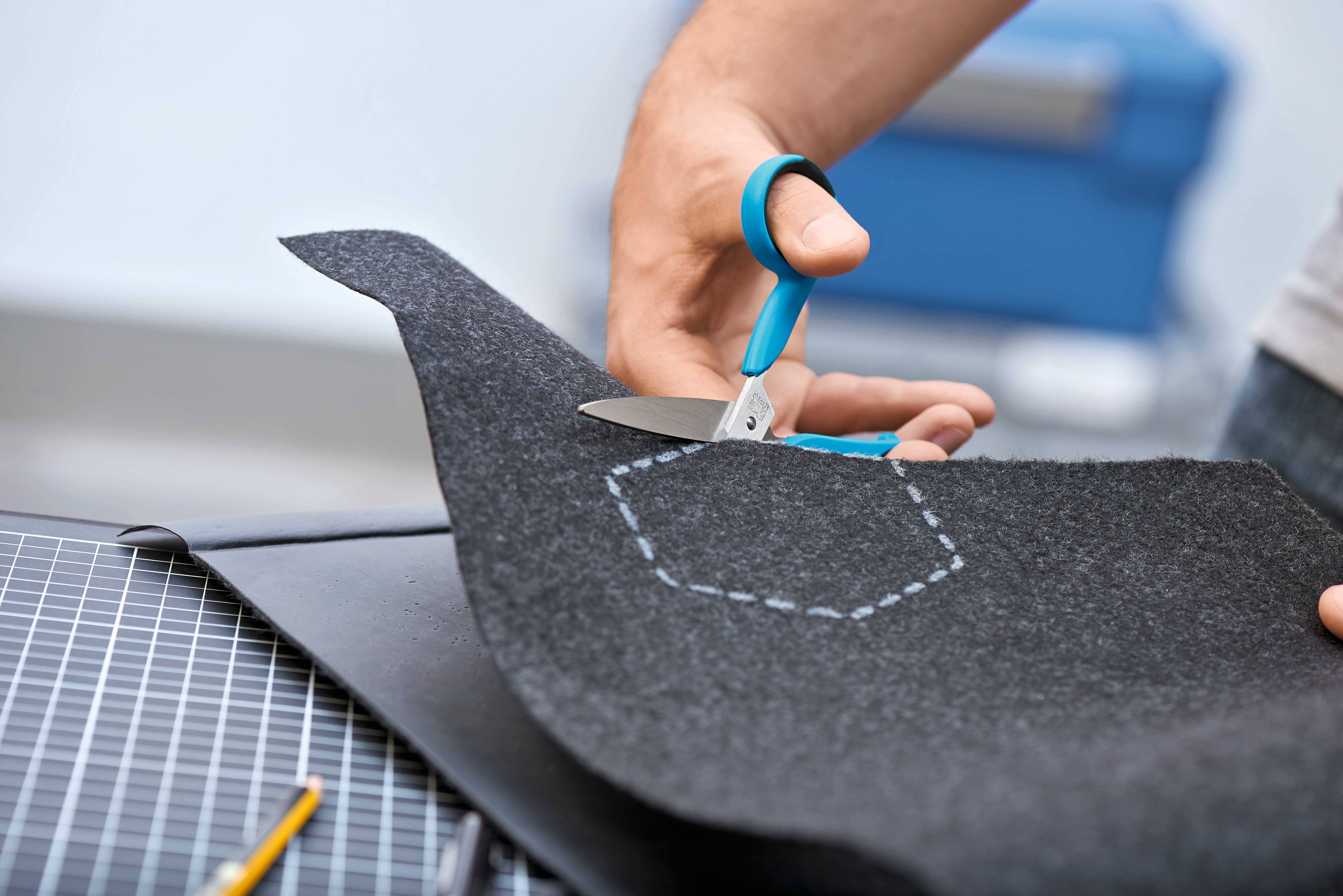 A hand holds a blue-green pair of scissors and cuts a grey fabric along a dotted line in a hexagonal shape.