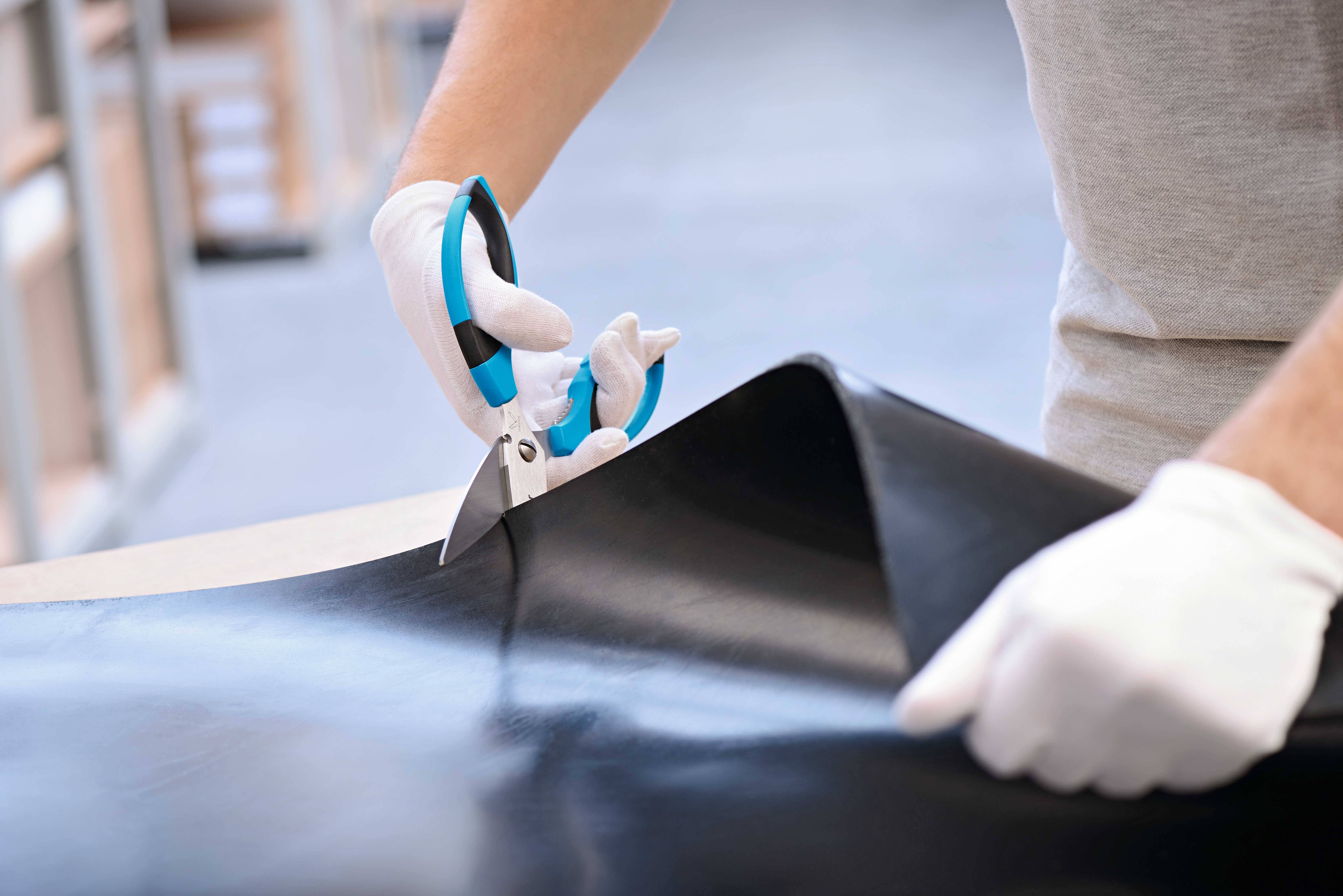 A person wearing white gloves is cutting a large black leather piece with blue scissors on a table.