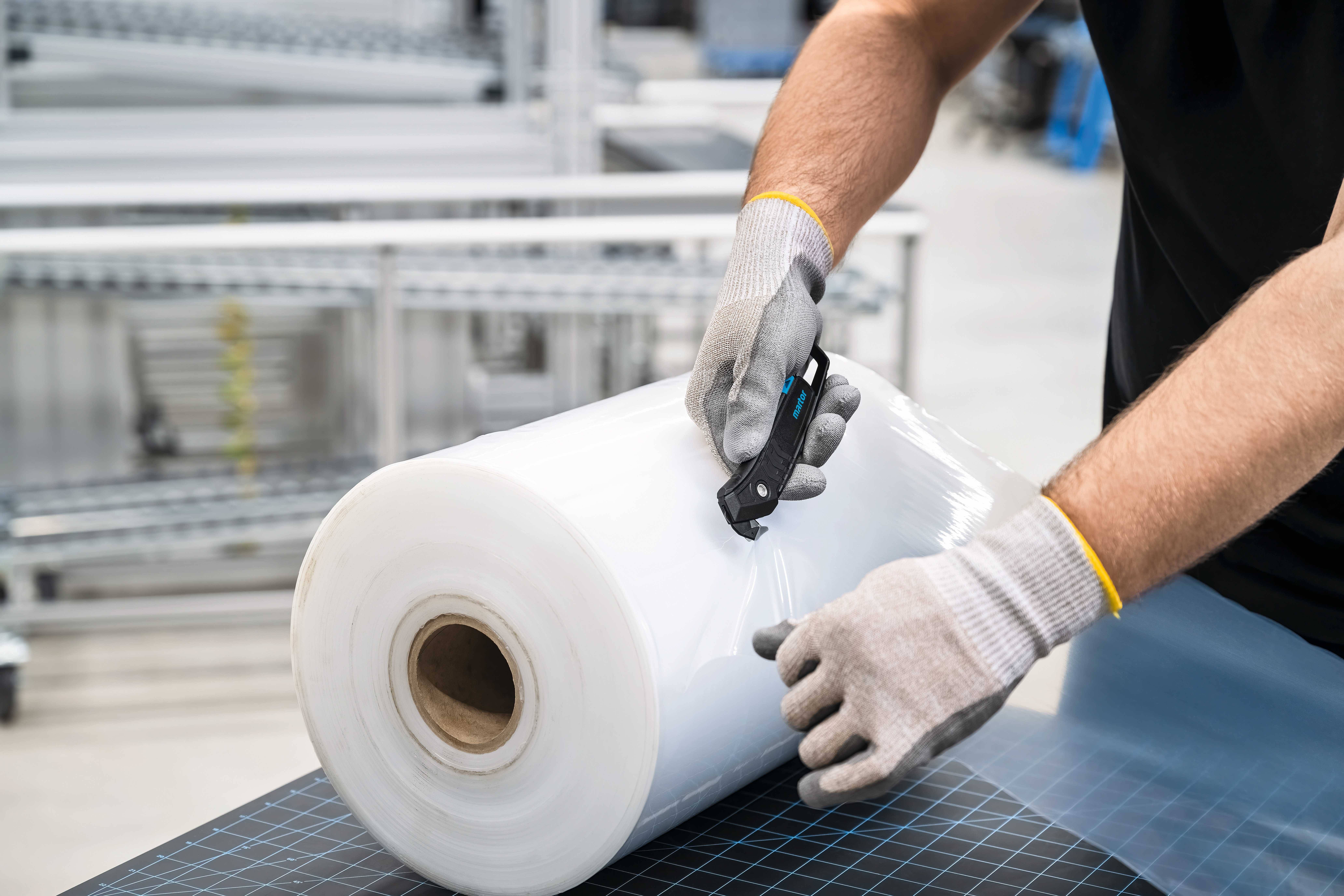 A person wearing gloves is cutting film from a large roll using a black knife. Shelves are visible in the background.