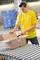 A man in a yellow T-shirt is opening a cardboard box on a conveyor belt in a warehouse. Stacked boxes can be seen in the background.