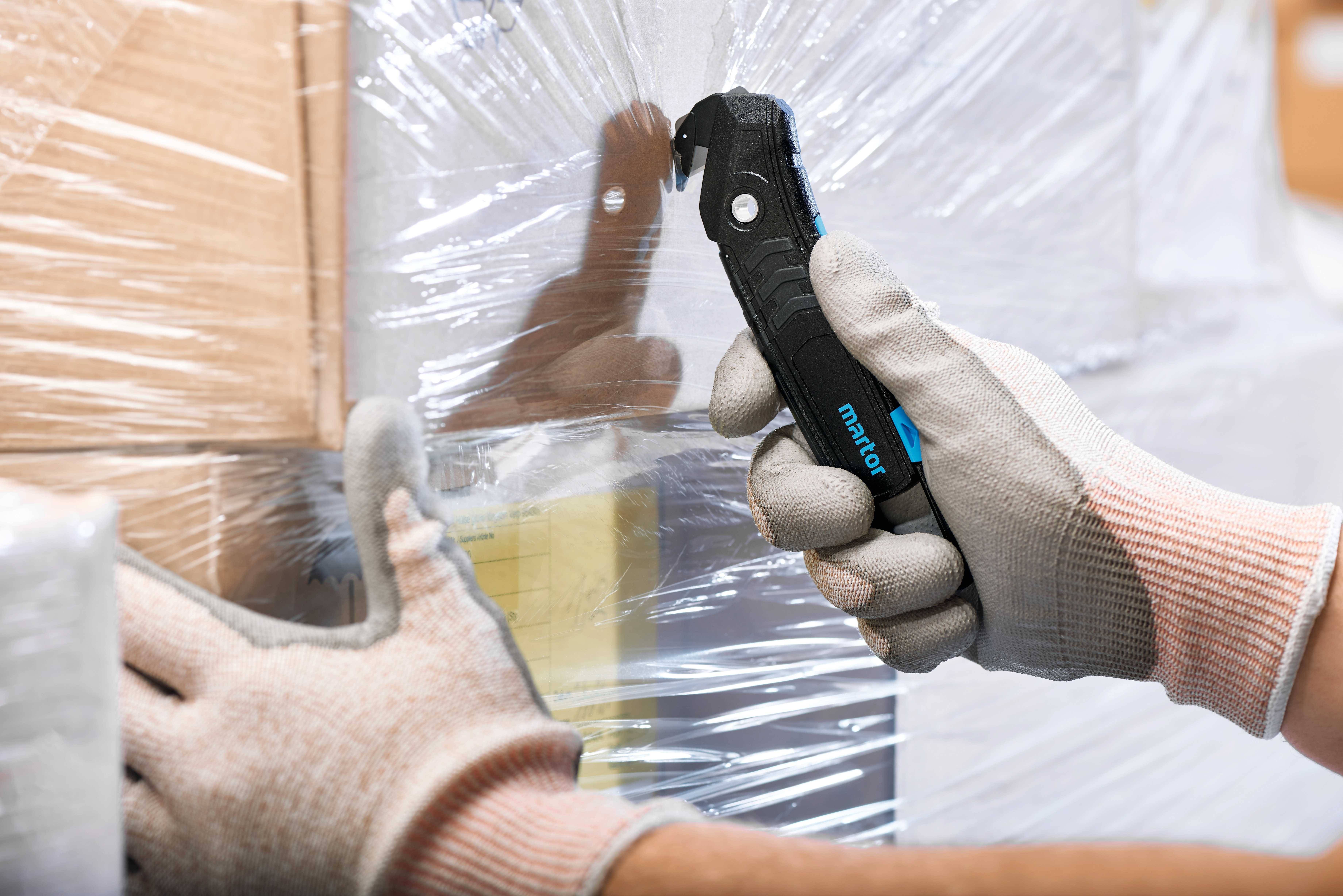 A hand uses a safety knife to cut packaging film from a stack of cardboard boxes. The person is wearing gloves for additional protection.
