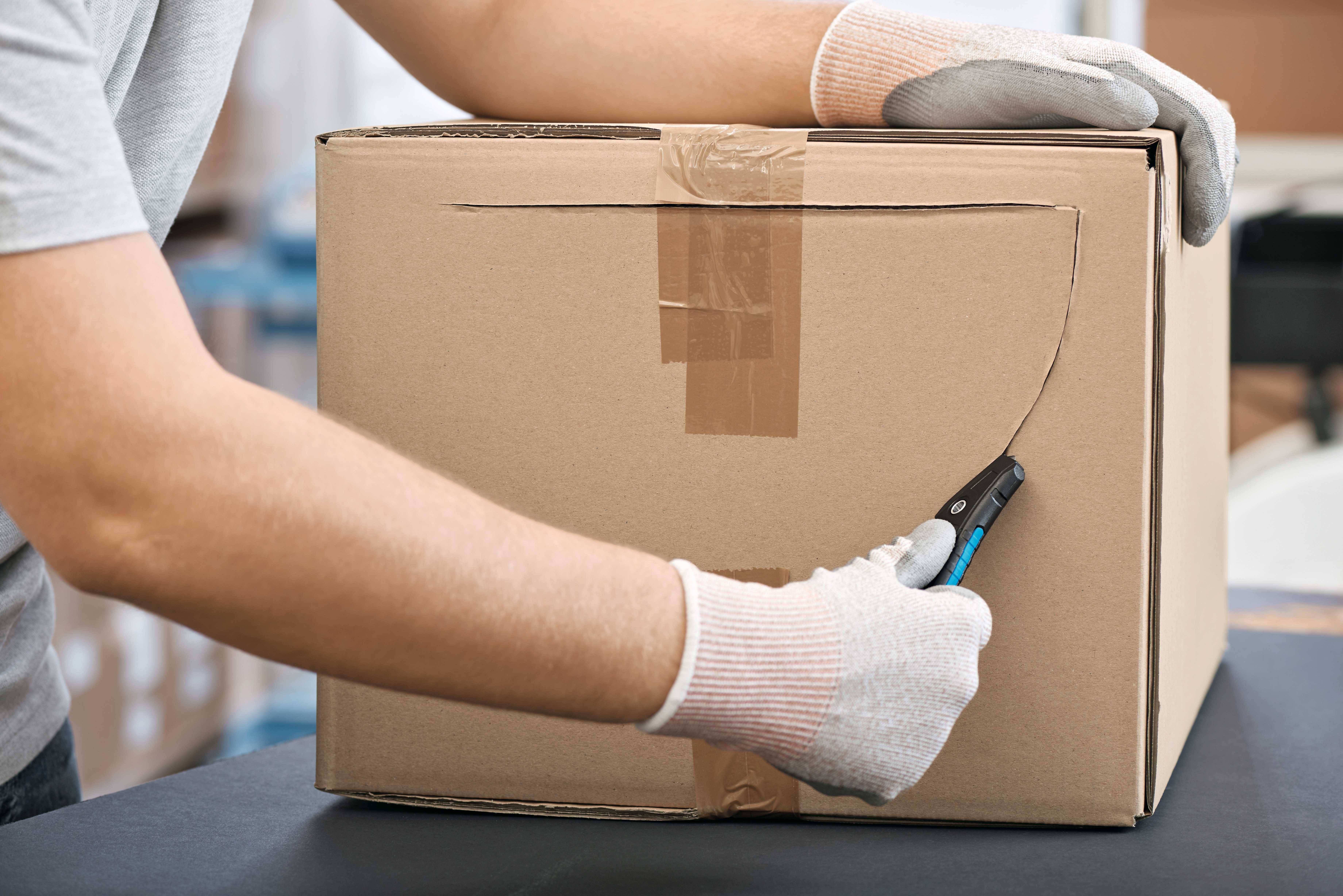 'A person carefully cuts open a cardboard box using a craft knife. Wearing protective gloves. Packaging is being opened.'
