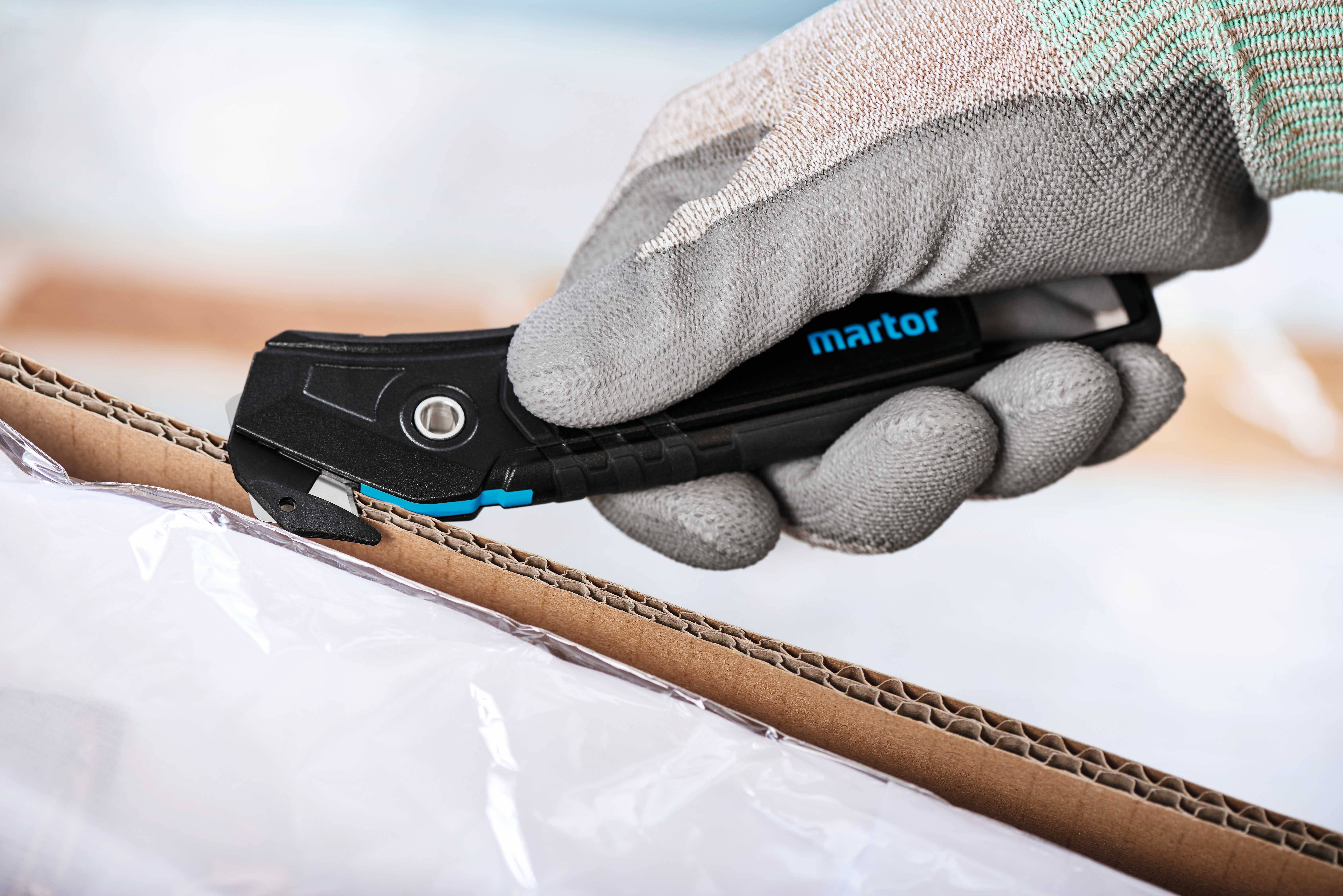 A gloved hand uses a black safety utility knife to open a cardboard box. The knife bears the 'martor' inscription.