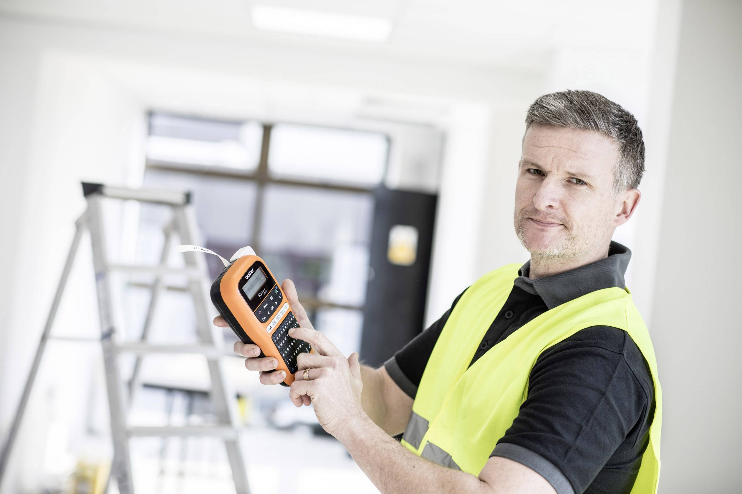 A man in a high-visibility vest is holding a device, possibly a measuring instrument, in a bright room with a ladder in the background.