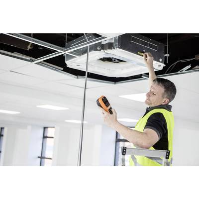 A technician wearing a high-visibility vest is checking the ceiling air conditioning system with a measuring device in an office.