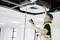 A technician wearing a high-visibility vest is checking the ceiling air conditioning system with a measuring device in an office.