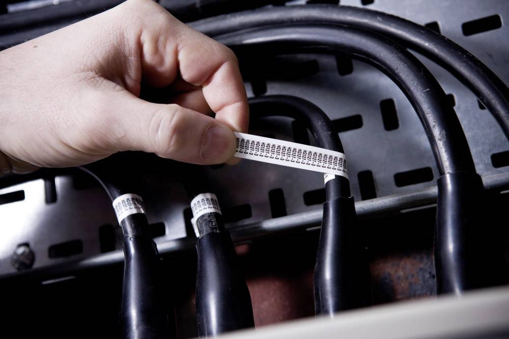 'A hand holds a label with a barcode, which is being attached to a black cable in a server or electrical area.'