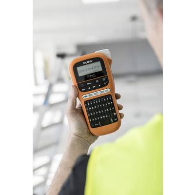 A person is holding an orange Brother label maker, with a blurry background featuring a ladder and work equipment.