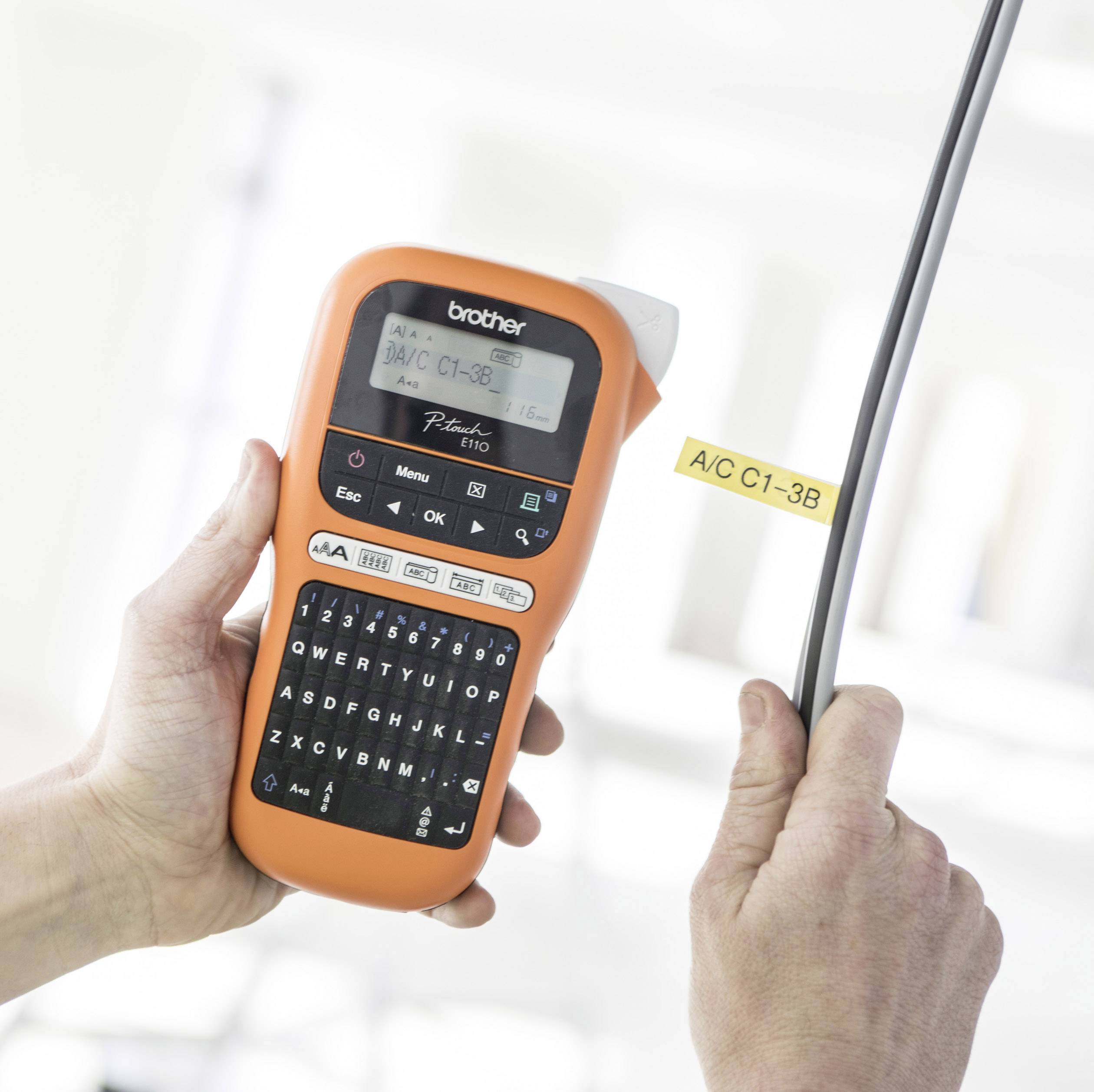 Holding an orange labelling device in hand. Display shows 'A/C C1-3B'. Label being applied to cable for identification.