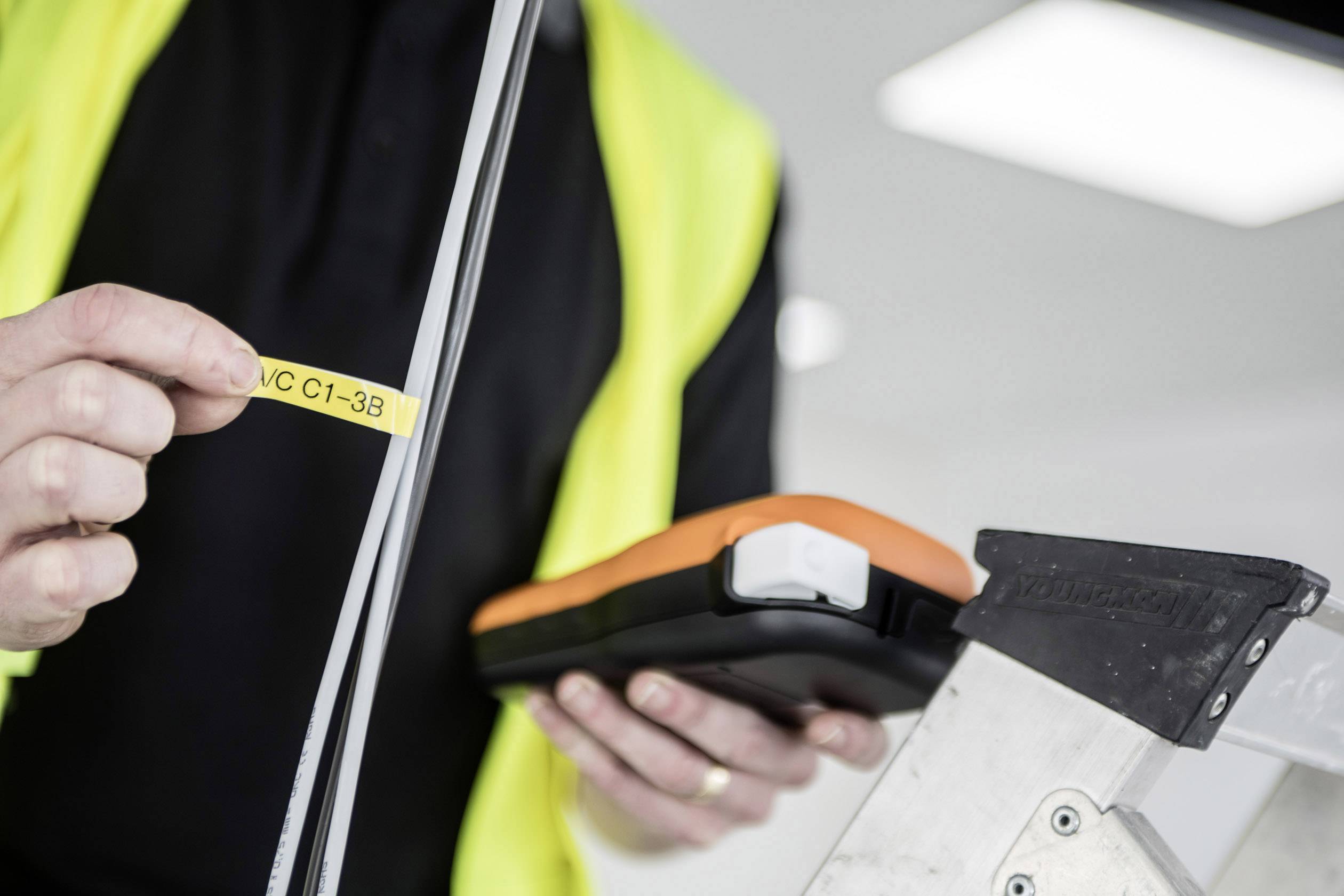 A person wearing a high-visibility yellow vest is holding an orange and black device and a yellow labelling strip next to a ladder.