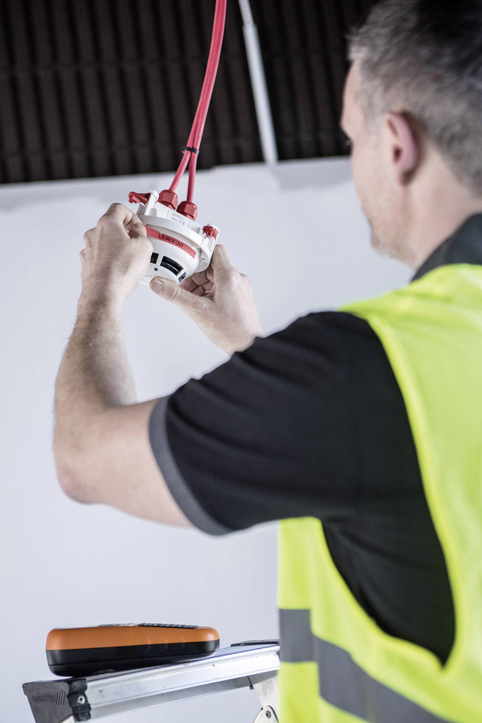 A construction worker in a yellow hi-vis vest is installing a smoke detector ceiling mount. He is standing on a ladder and holding red cables.