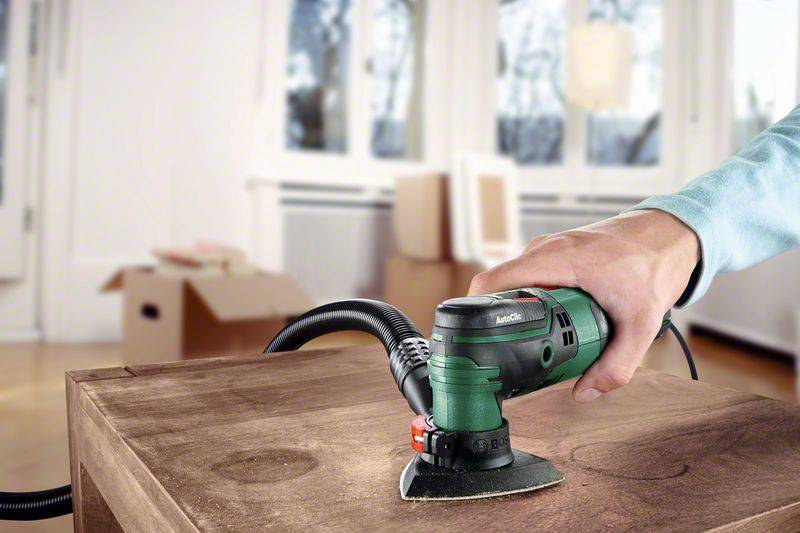 A person is sanding a wooden surface with an electric sander. Cardboard boxes and a window are visible in the background, suggesting a renovation context.