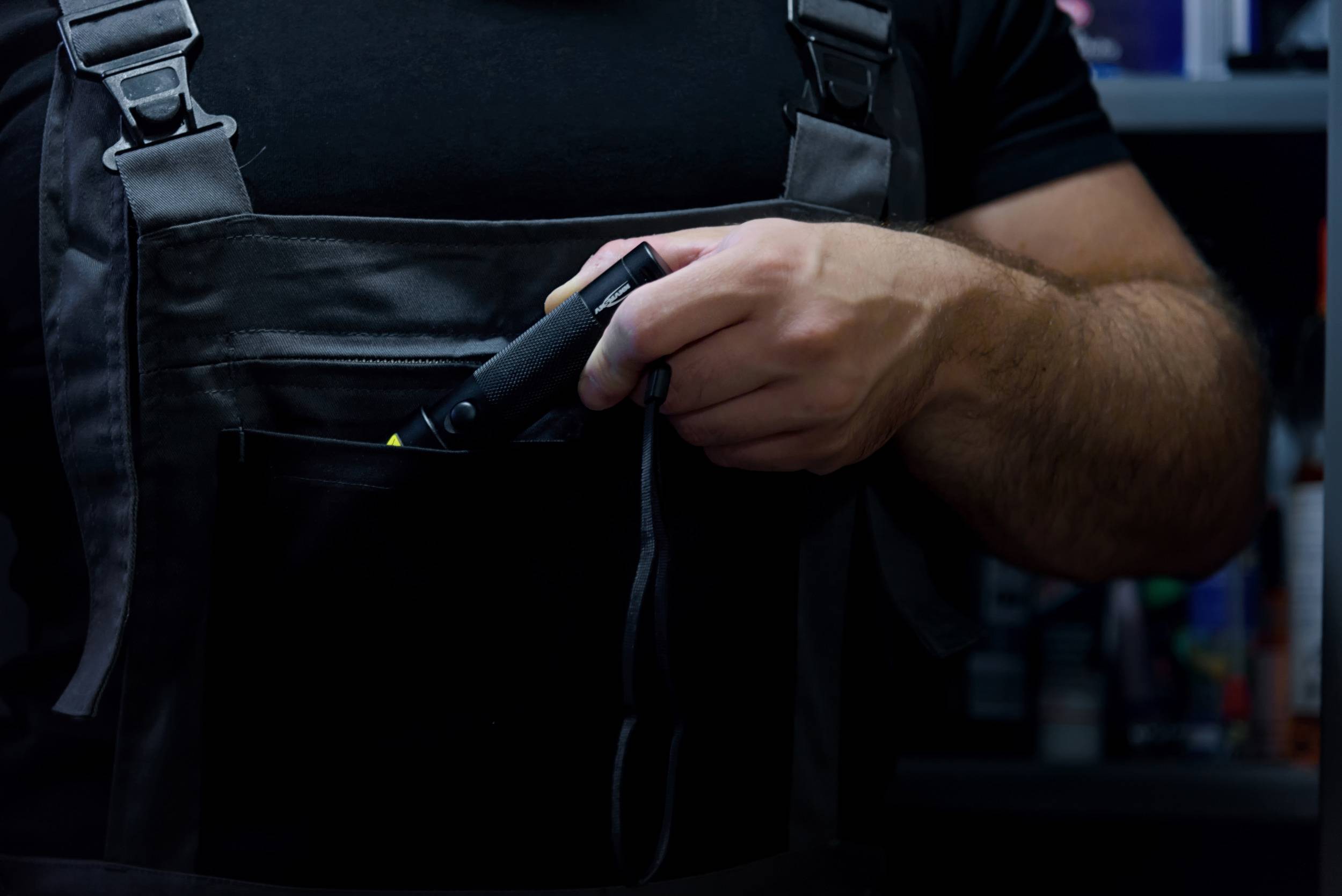 A person is placing a tool in the pocket of their work dungarees. They are standing in front of a blurred background with shelves.