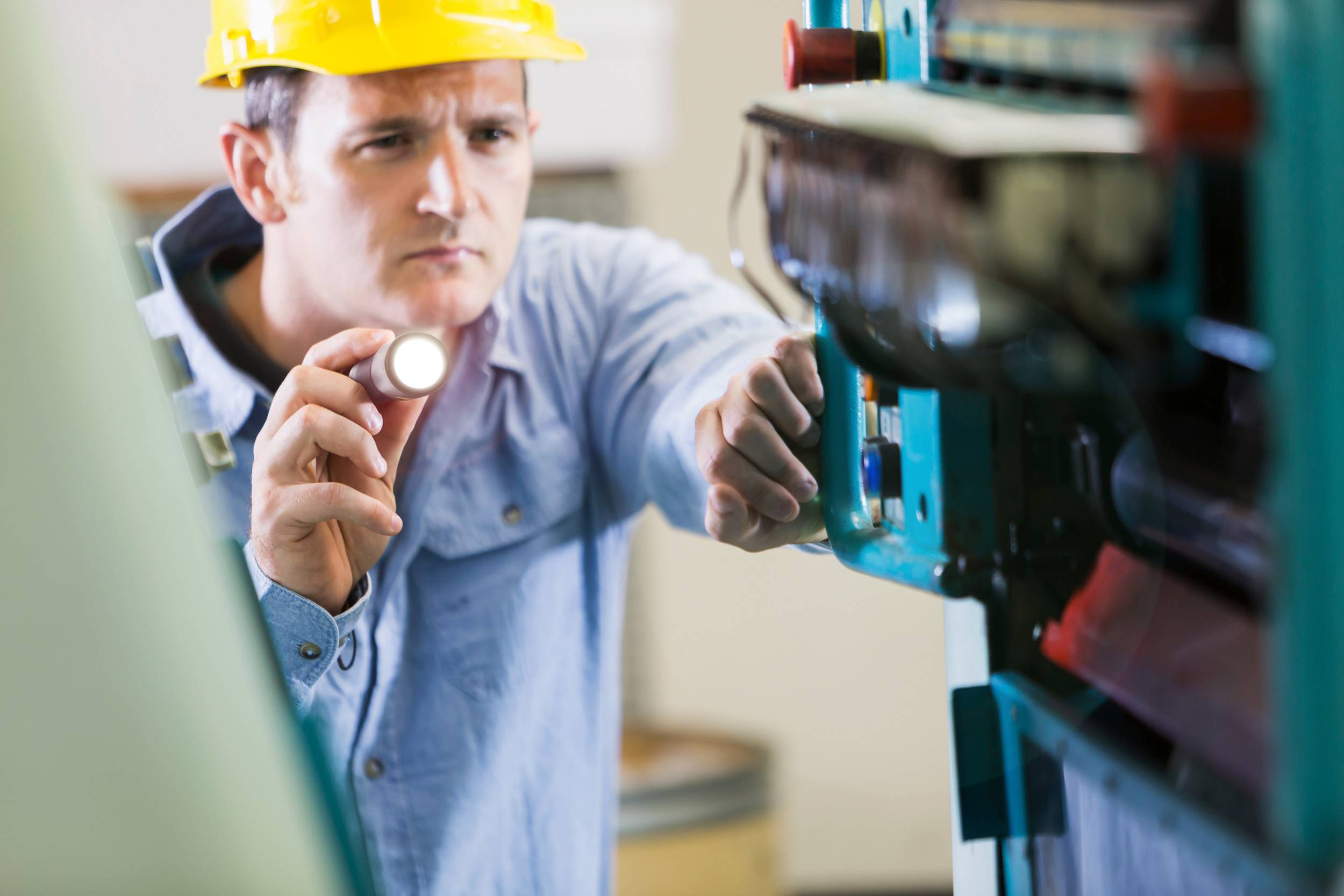 A man wearing a hard hat is inspecting a machine with a torch. He is wearing a blue shirt and is focusing intently on the equipment.