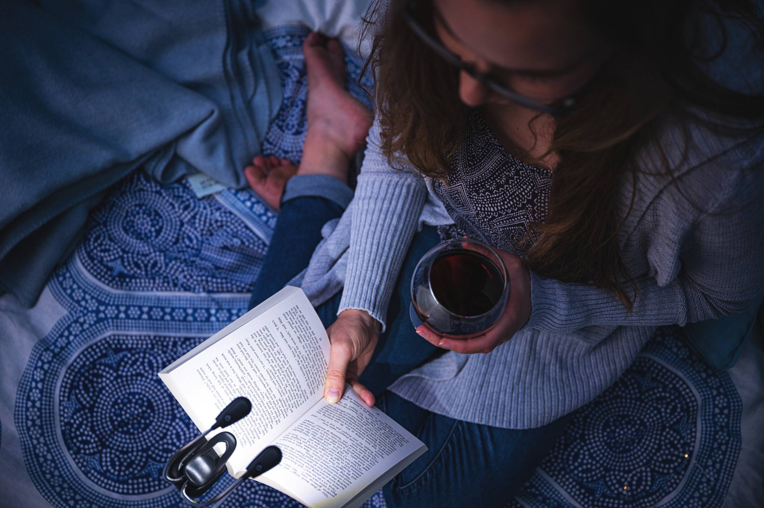 A person is reading a book on a blanket, holding a drink in their other hand. They appear relaxed in a cosy atmosphere.