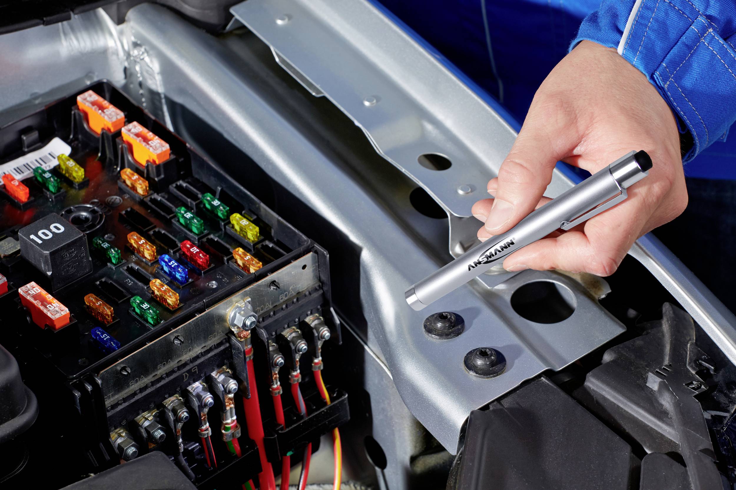 A hand is holding a voltage tester over an open fuse box in a car's engine compartment. Various coloured fuses are visible.