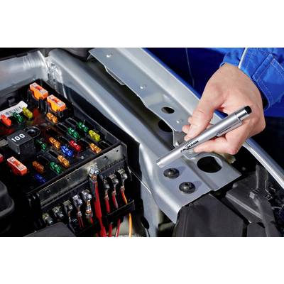 A hand is holding a voltage tester over an open fuse box in a car's engine compartment. Various coloured fuses are visible.