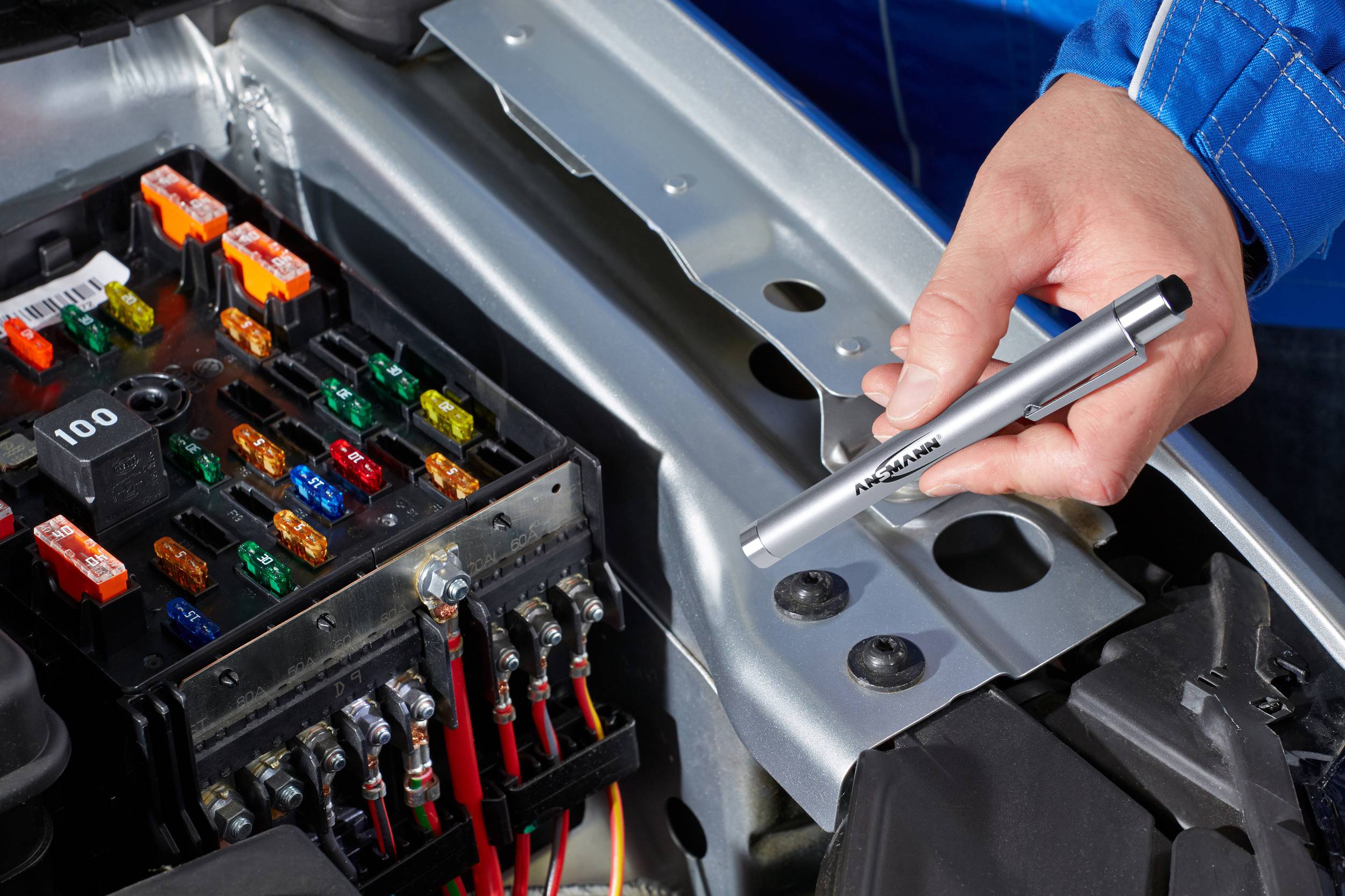 A person is holding a multimeter probe over an open fuse box of a car. Various coloured fuses are visible.