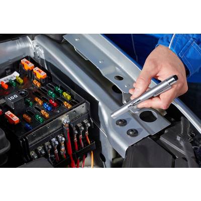 A person is holding a multimeter probe over an open fuse box of a car. Various coloured fuses are visible.