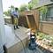 A man is cleaning a tiled patio with a pressure washer. Next to him stands the machine, surrounded by plants and fences.