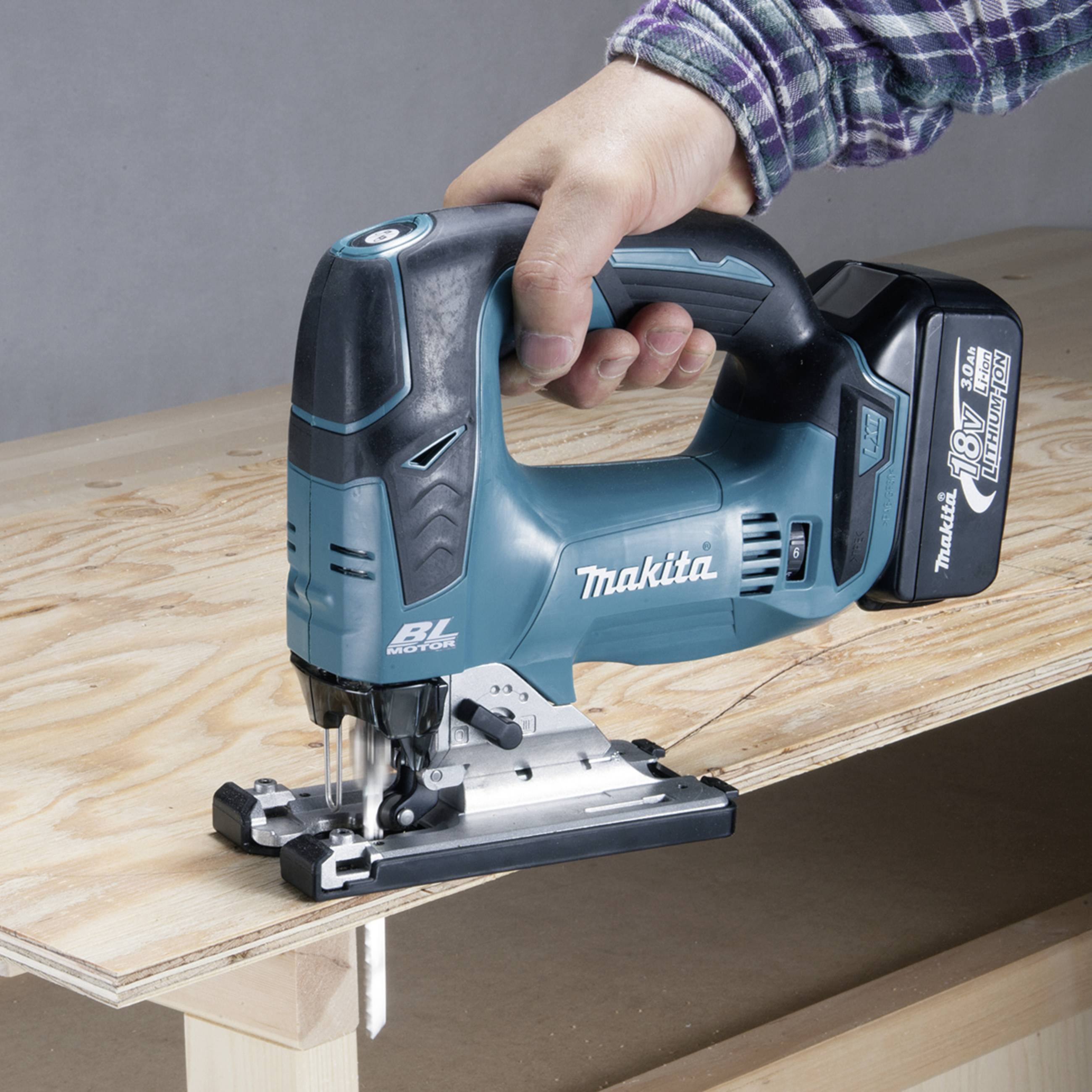 A hand is holding a blue jigsaw, which is about to cut a piece of plywood. The saw is cordless and ready for use.