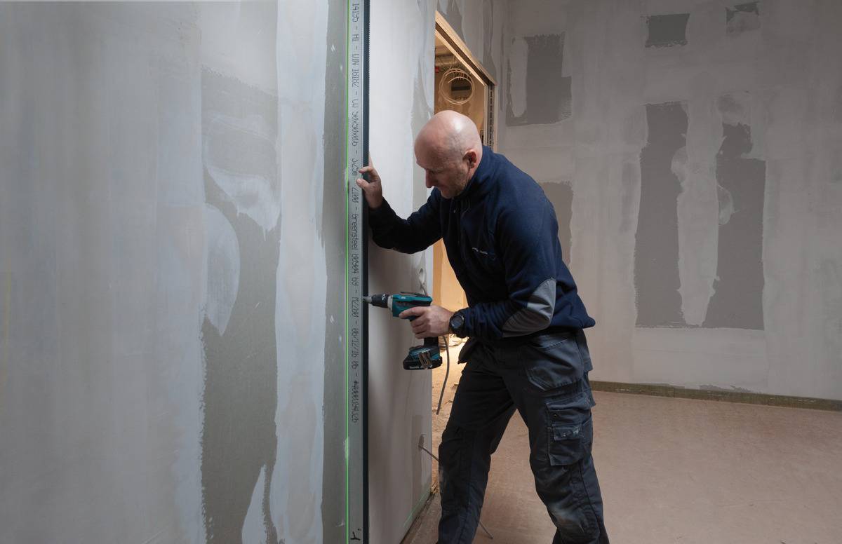 A tradesman is using a drill to secure a long ruler to a wall. The wall is partially plastered.