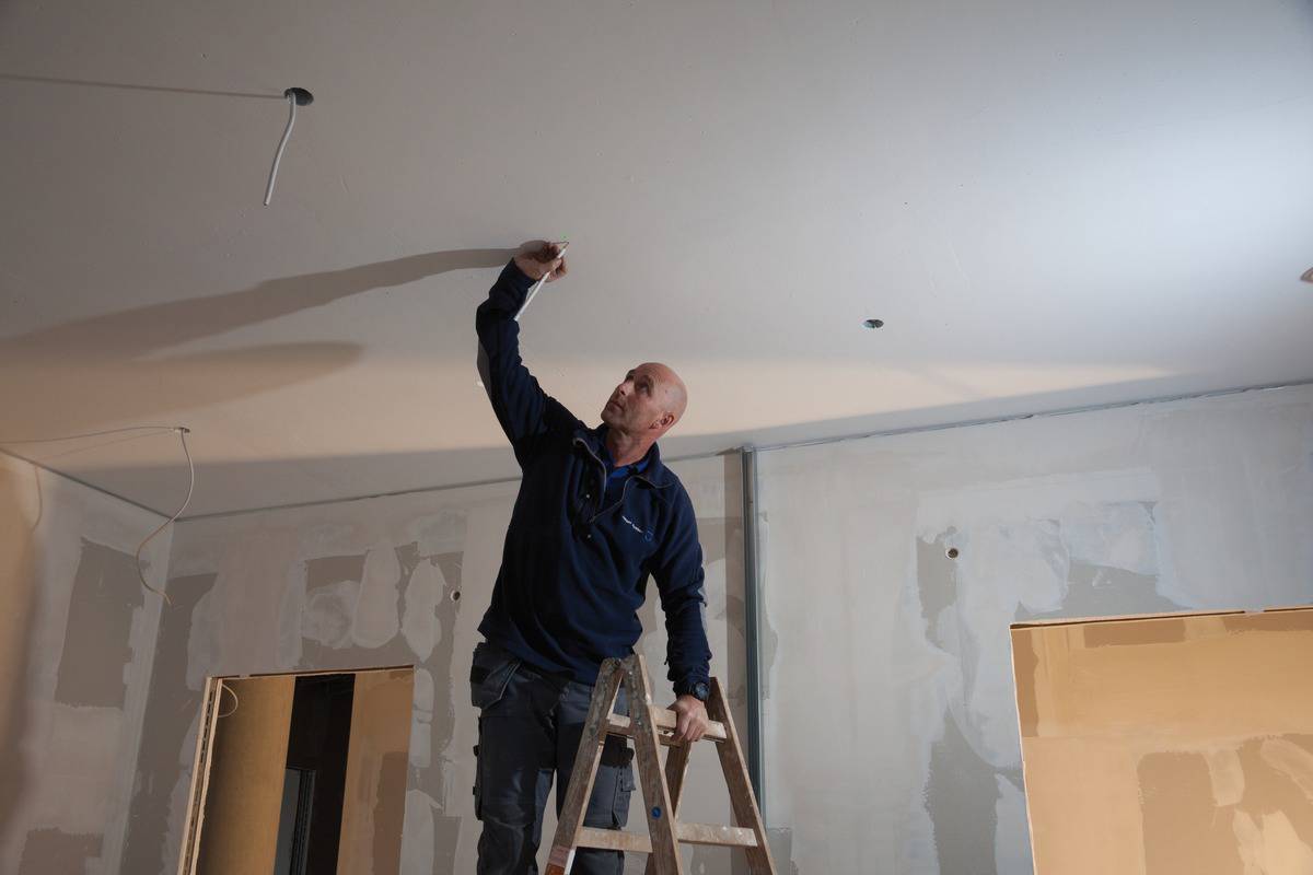 A man is standing on a ladder and working on the ceiling of an unfinished room. Walls are partially plastered.